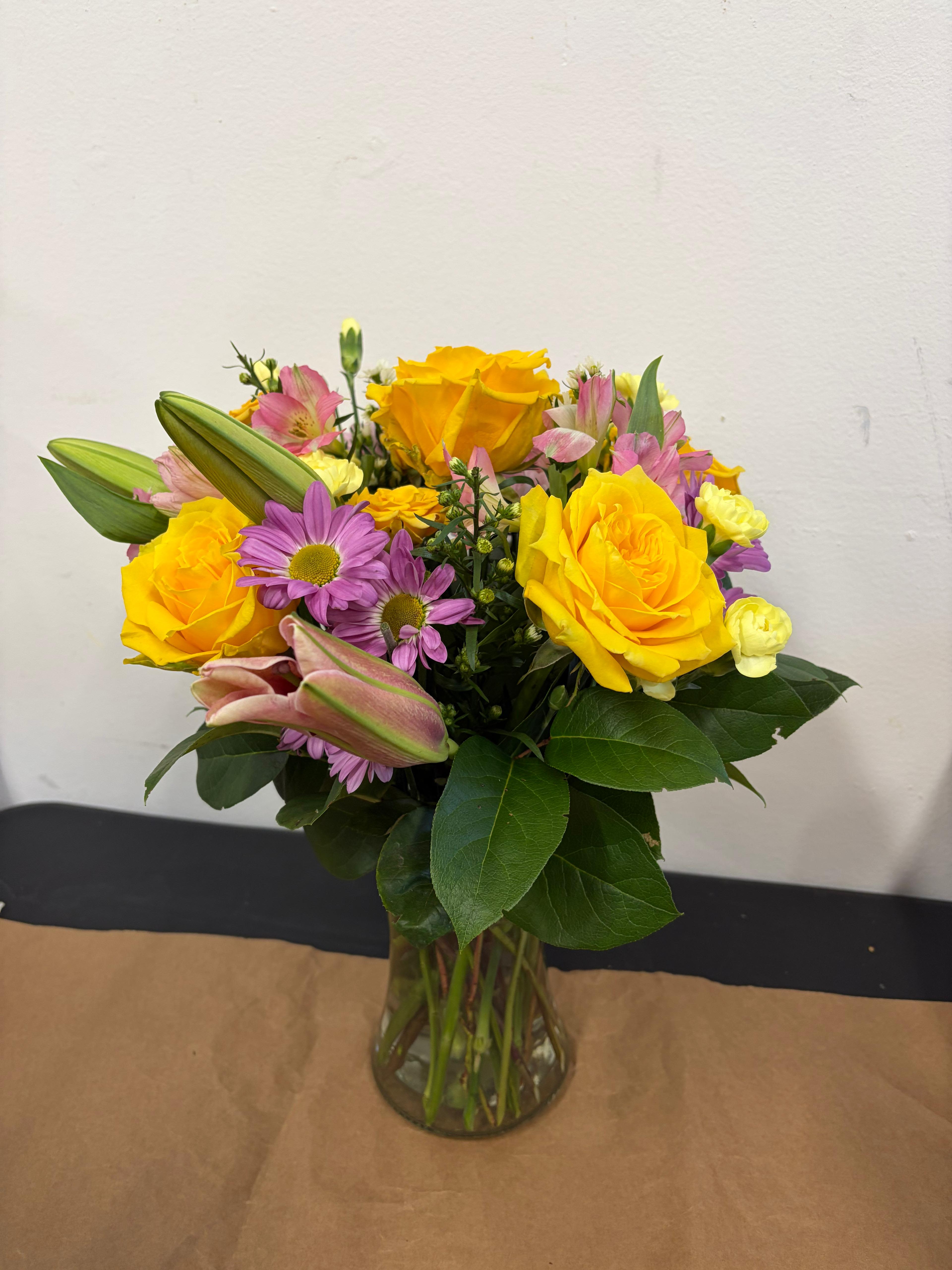 Sun-Drenched Serenity  - Brighten any room with the vibrant and uplifting Sun-Drenched Serenity. This cheerful arrangement centers around bold, sunny yellow roses that radiate warmth and happiness. Complemented by the delicate, star-shaped petals of purple daisies and the exotic elegance of pink alstroemeria, this bouquet is a masterpiece of color and texture. Housed in a classic clear glass gathering vase, it’s an ideal gift for birthdays, "get well" wishes, or simply to bring a touch of a summer garden indoors. 