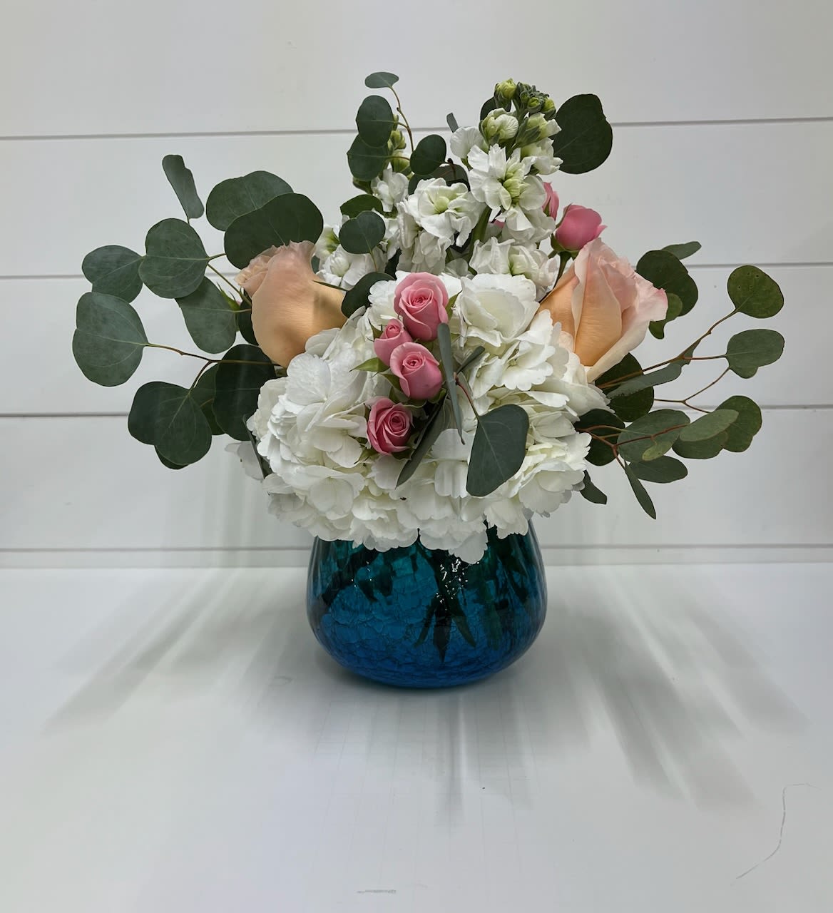 Gentle Spring - Celebrate the softness of the season with Gentle Spring, a graceful floral arrangement designed to bring calm beauty into any space. This elegant design features lush hydrangea, classic roses, and delicate spray roses, thoughtfully accented with fresh, airy greenery for a naturally refined look.  A soothing palette of peach, pink, and crisp white blooms creates a romantic, garden-inspired feel, while the blue and green vase adds a refreshing pop of color—reminiscent of clear skies and new spring growth. The combination feels both timeless and uplifting, making it perfect for Easter, spring gatherings, or simply brightening someone’s day.  Gentle Spring is a beautiful expression of renewal, warmth, and quiet elegance—ideal for gifting or as a centerpiece that brings the season indoors.  Delivery Available In: Smyrna, Marietta, Mableton, Austell, and Atlanta.  Substitution Policy: To ensure the freshest arrangement possible, flower varieties or vase styles may be substituted based on seasonal availability while maintaining the overall look, color palette, and value of the design.