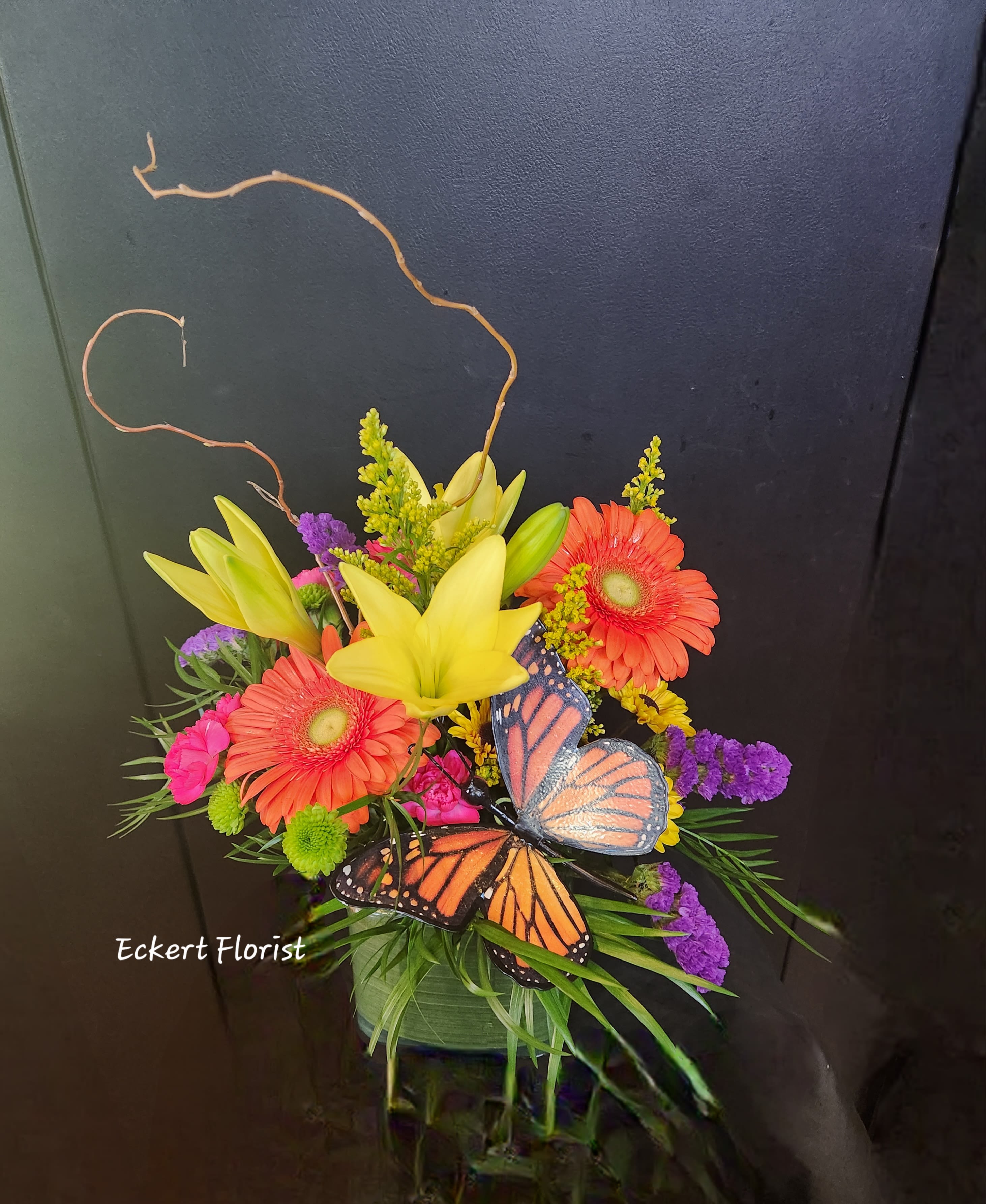 Eckert Florist's Butterfly Kisses Bouquet *Our Local Delivery Only - This vibrant mix of lilies, gerbera, carnations and mums in a leaf-wrapped glass vase with a keepsake monarch butterfly pick and curly willow will sure to brighten someone's day. Flower varieties and colors may vary. Our local delivery only.