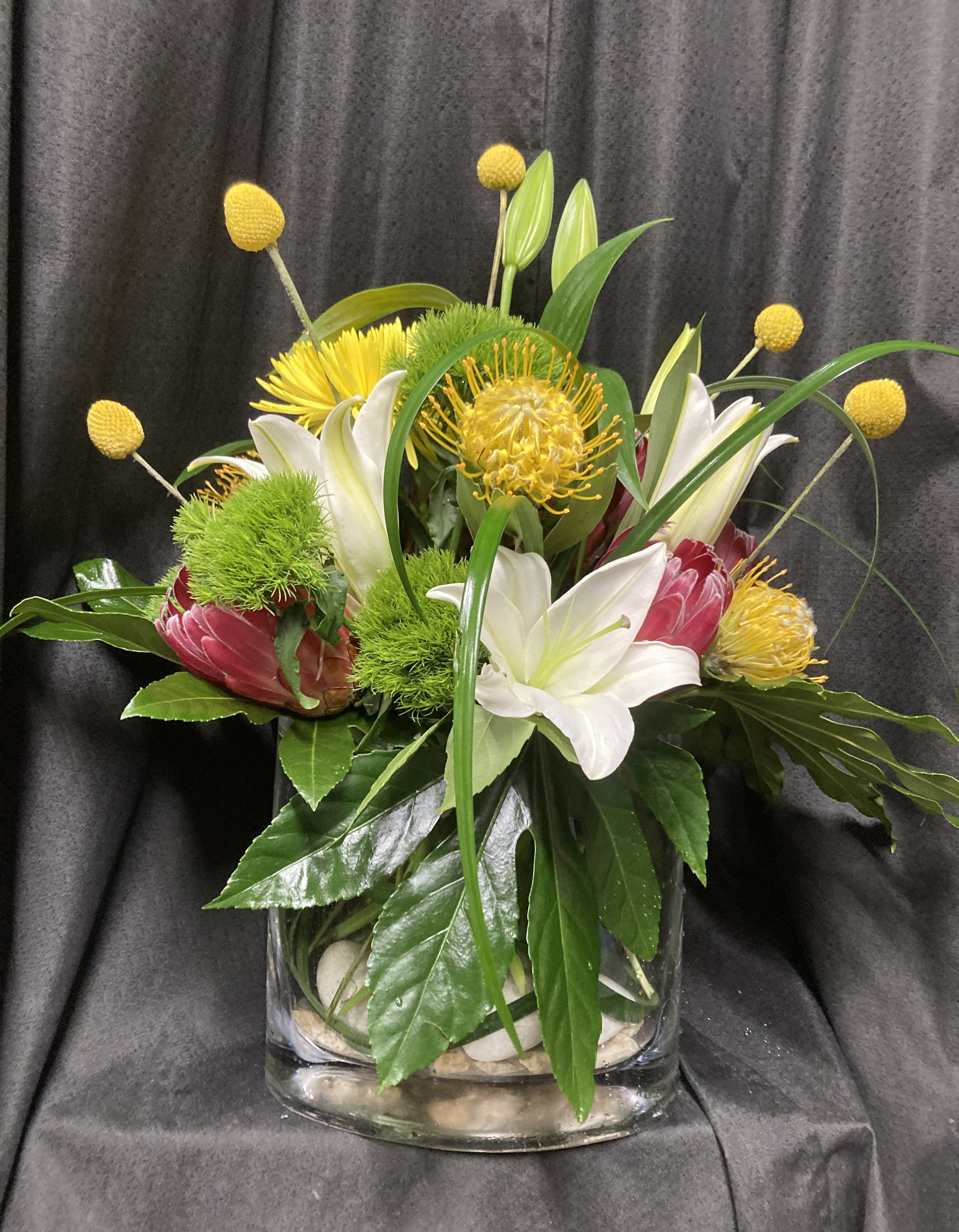 TROPICAL BEAUTY - ASSORTED TROPICAL BLOOMS AND GREENS IN A CUSTOM TOP SHELF VASE.