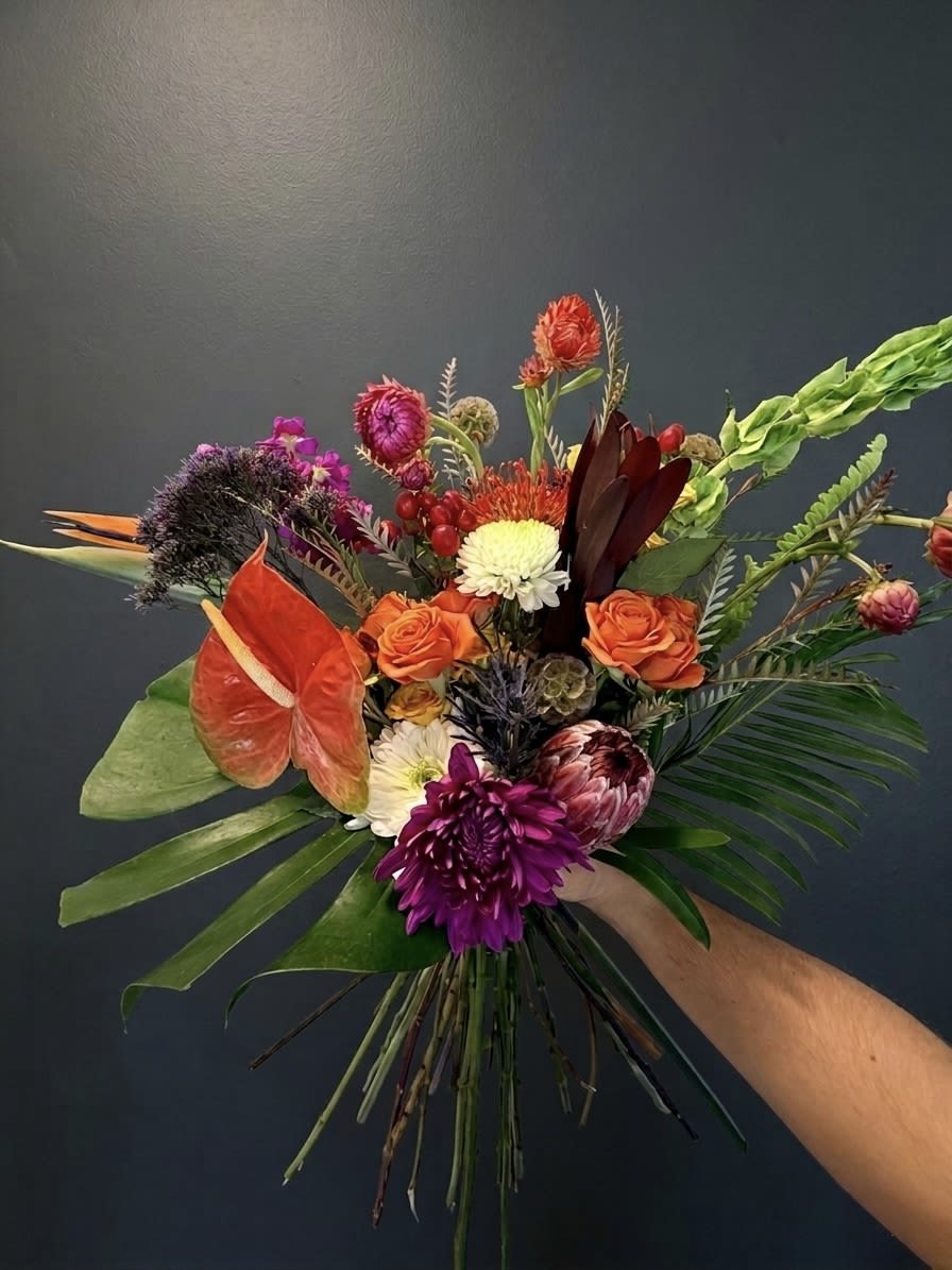 Tropic No. 1 - A masterclass in texture and tonal contrast, Tropic No. 1 is a living sculpture designed for the botanical maximalist. This curated arrangement juxtaposes the structural defiance of Birds of Paradise, Anthurium, and King Protea against the velvet-soft ruffles of Saffron Spray Roses and Lavender Trachelium. Inspired by the high-contrast beauty of a tropical sunset, we’ve layered an architectural base of Monstera and Sword Fern with the glowing embers of Red Pin Cushion Proteas and the intricate curiosities of Scabiosa Pods and Blue Thistle. It is a bold, premium statement piece that brings a lush, high-end escape into the modern home.