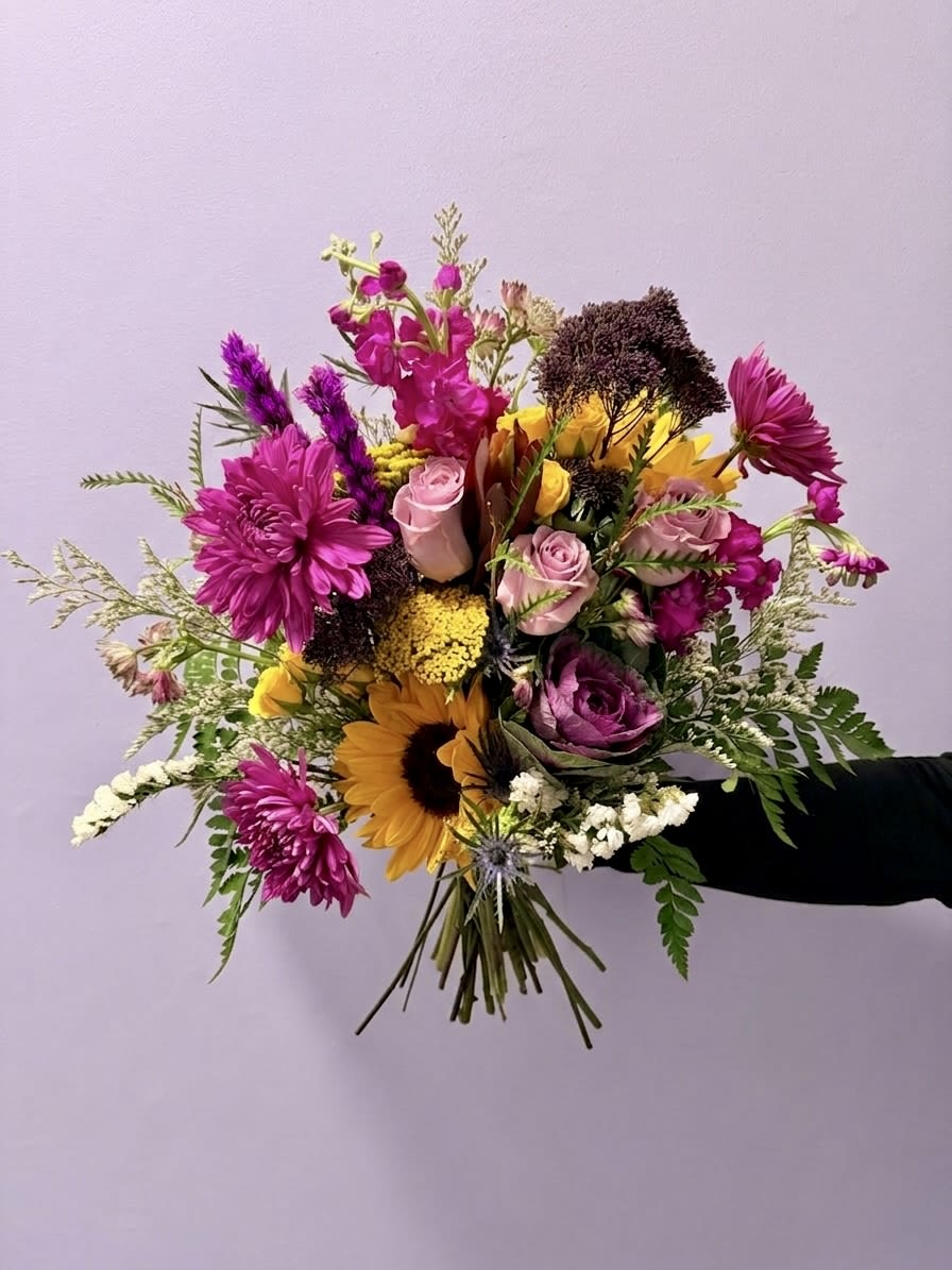 Amethyst Meadow  - A vibrant, textured arrangement that captures the wild elegance of a Southern garden at twilight. This curated piece balances the deep, moody purples of ornamental kale, trachelium, and liatris against the sun-drenched gold of sunflowers and yarrow. Architectural safari protea and airy astrantia add modern sophistication, while plush cremone, stock, and a variety of purple roses provide a timeless, romantic finish.