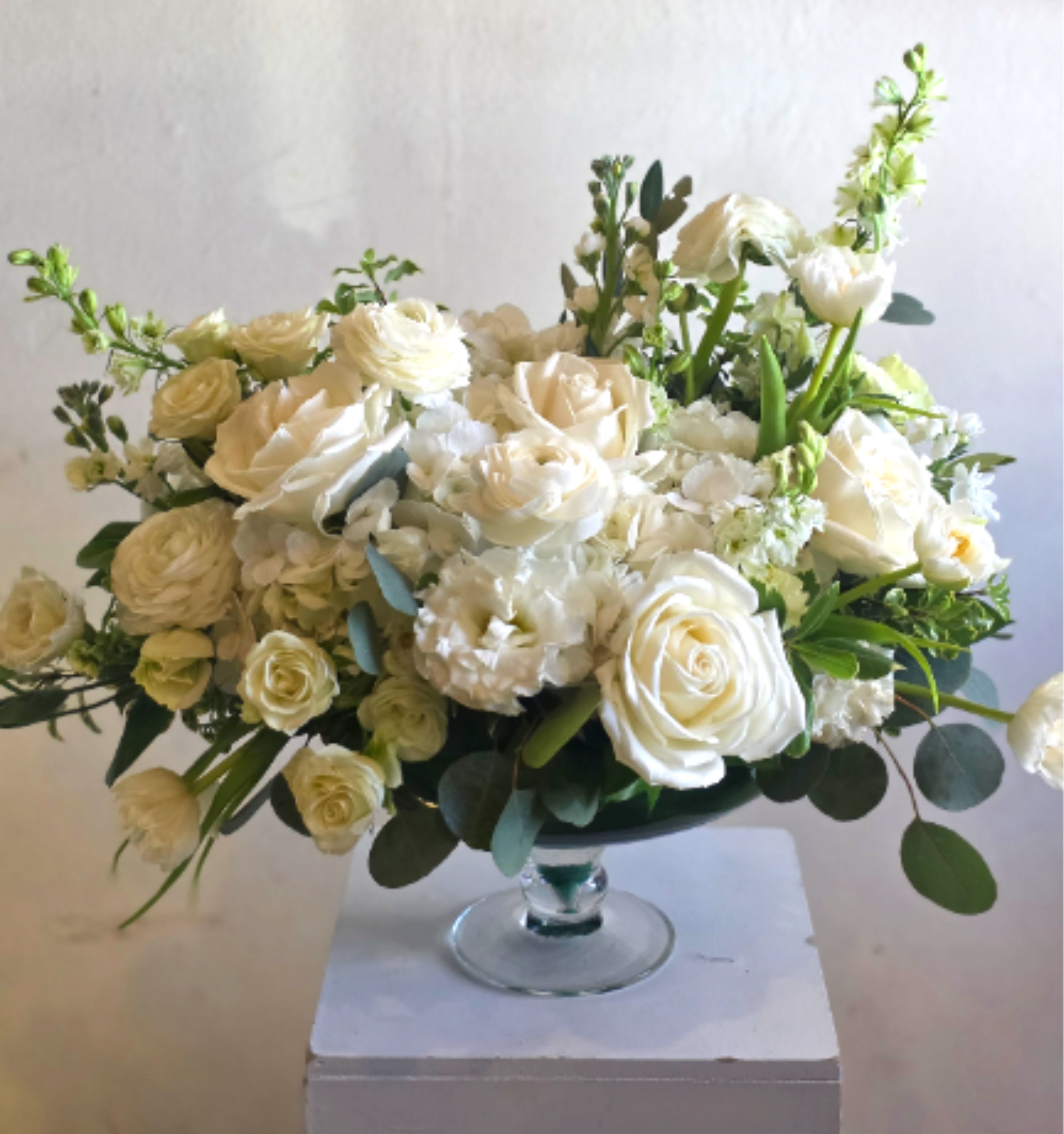White Garden Luxe - A cloud of creamy whites-white roses, ranunculus, lisianthus, hydrangeas, and stock-forms a refined tabletop centerpiece perfect for a wedding centerpiece, anniversary bouquet, or an elegant sympathy arrangement. The low glass pedestal bowl keeps the design intimate and polished, so the person you're thinking of will feel comforted and truly seen by its serene, sophisticated presence.  Flowers are densely clustered for a soft, rounded silhouette with subtle shifts in ivory and green foliage tucked throughout for fresh contrast. Taller stems at the back and sides introduce gentle movement while the wide, shallow shape makes it ideal for dining tables, reception rounds, or a mantel display. Delivered in a clear glass pedestal bowl, this arrangement is sized for easy placement and instant impact; refresh water as needed to keep blooms at their best.