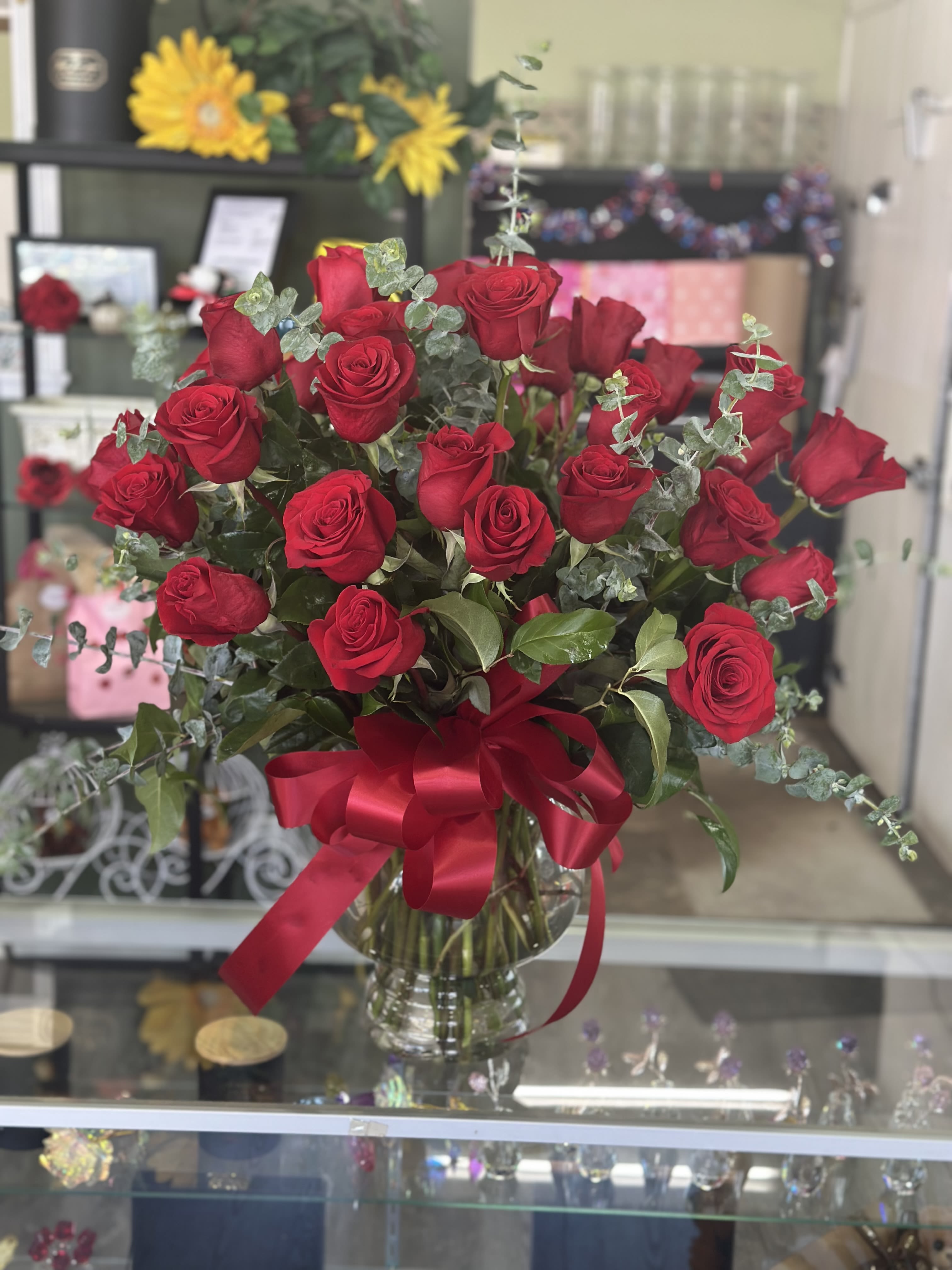 24 Long stem roses  - A generous bouquet of 24 long-stem red roses gathered in a flared clear glass vase creates an immediate impression of romance and presence-perfect as an anniversary bouquet or a bold birthday surprise. The rich crimson roses are arranged in a full, rounded profile with a few stems rising slightly for natural movement, giving the person you're thinking of a dramatic yet elegant gift that says "I love you," "I'm sorry," or simply "you matter" without words.  Textural filler and cool-toned accent foliage are woven through the blooms to add contrast and volume while keeping the red roses front and center. The arrangement sits in an 11" clear vase and is finished with a wide red satin ribbon and a generous bow at the neck for a polished, gift-ready presentation. Ideal for love and romance, Valentine's Day, or thoughtful "thinking of you" moments, these long stems arrive ready to display and delight.