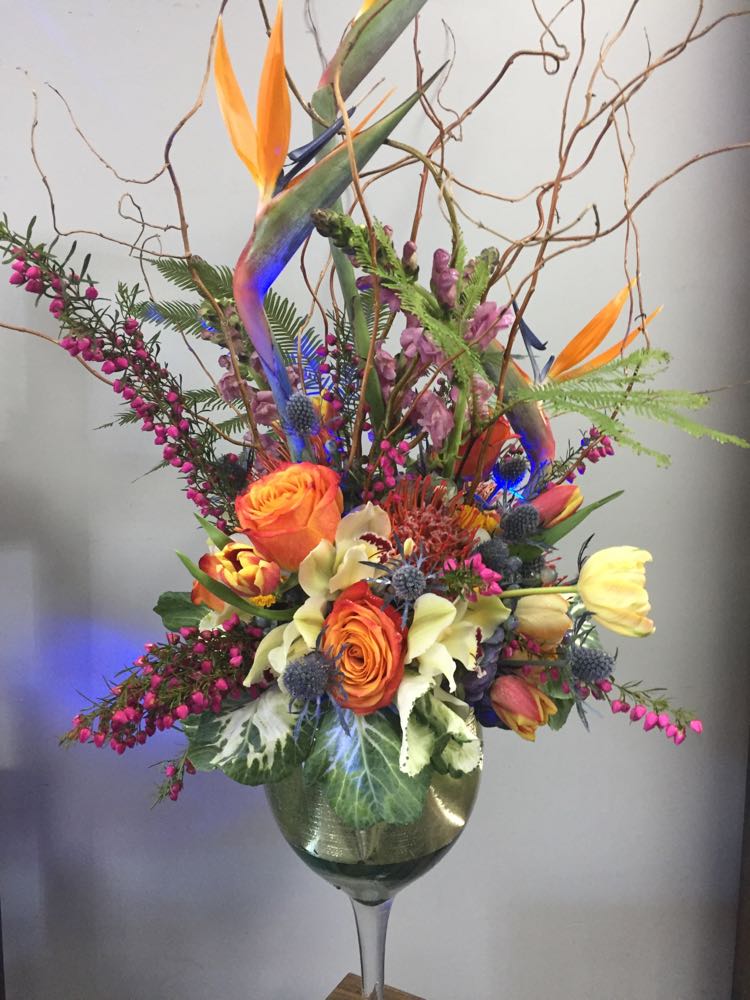 BLUE BIRDS - This elegant arrangement is filled with birds of paradise, tulips, kale, roses, snapdragons and curly willow. It is filled with assorted greens , designed in a tall cocktail vase, and accented with submersible blue lights. Unique and Elegant.