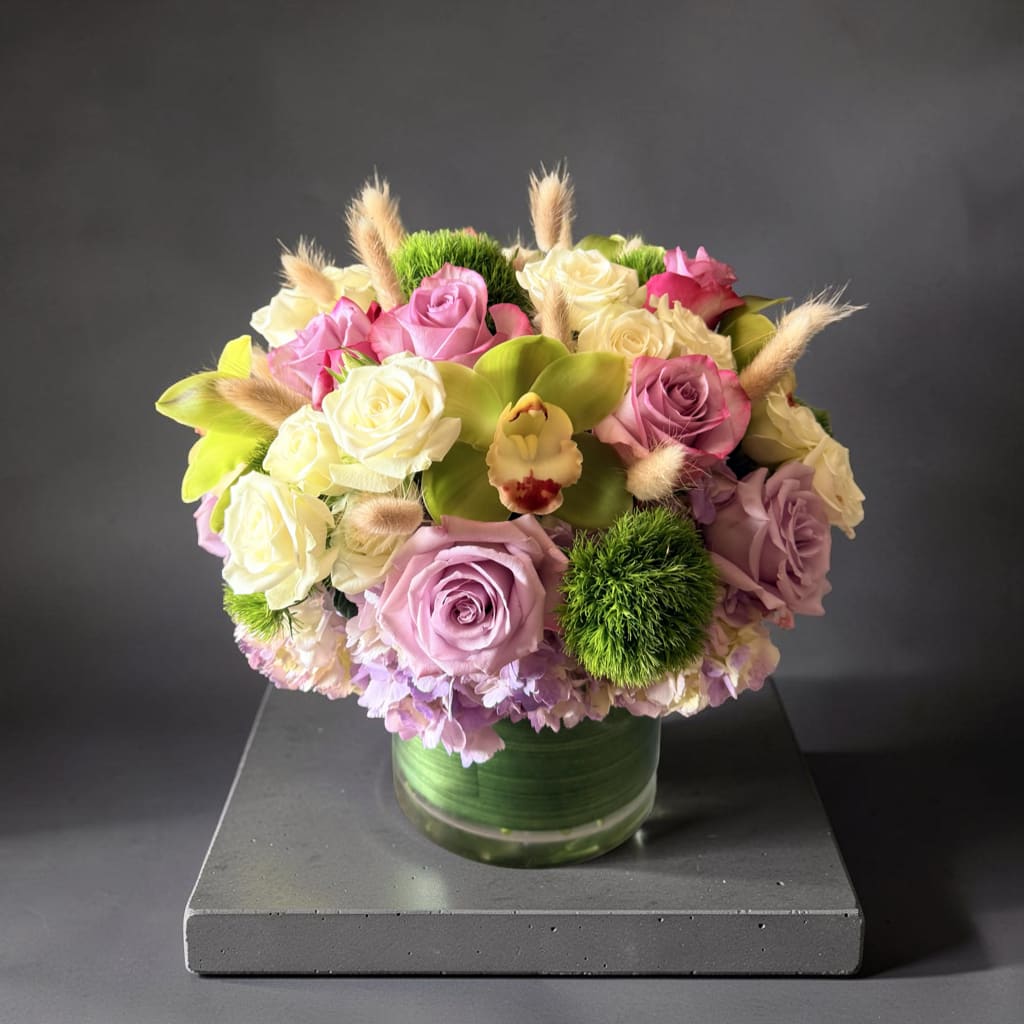 Blushing Orchid Garden - A soft and elegant arrangement featuring blush and ivory roses, a striking green cymbidium orchid, lush hydrangea, and textured green dianthus with delicate natural accents. Designed in a modern glass vase, this fresh garden-style piece brings charm, color, and sophistication to any space.(In 6x6 vase, great size for office meetings and small parties )