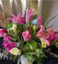 Pink lilies, roses, and pale calla lilies arranged with bright filler flowers in a white vase