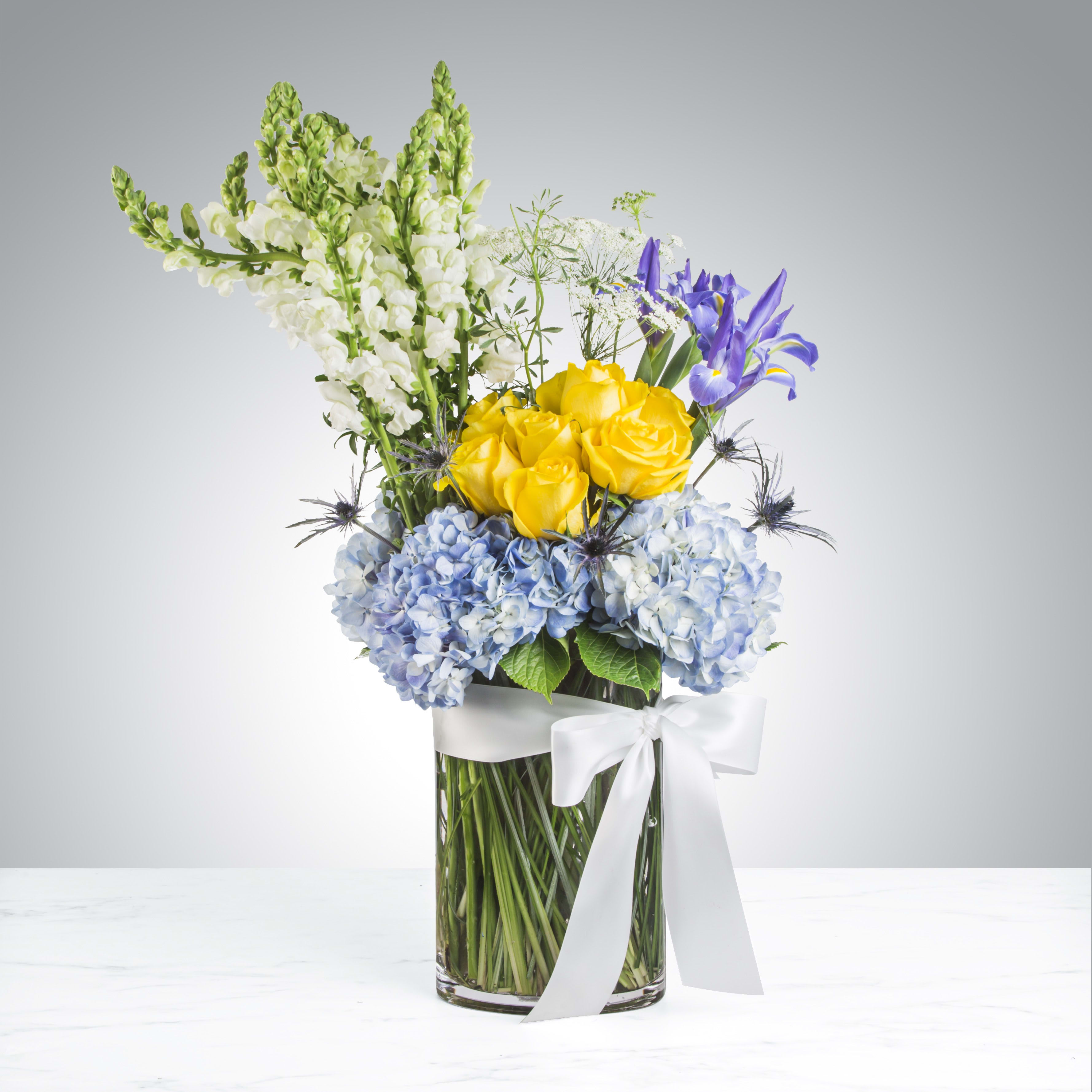 Yellow roses, blue hydrangeas, and purple irises with white snapdragons in a glass vase with white bow.