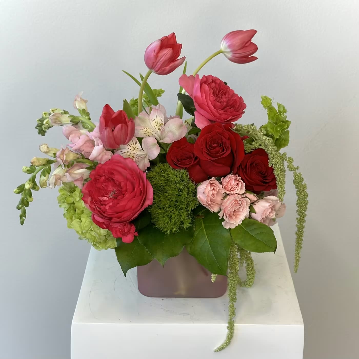 Strawberry Matcha - The Strawberry Matcha flower arrangement is a bold, playful, and ultra-luxurious design that blends vibrant pink tones with fresh green accents for a look that feels both trendy and unforgettable. Inspired by the popular flavor pairing, this arrangement brings together a delicious mix of color, texture, and movement, making it a standout choice for birthdays, celebrations, or statement gifting.  Rich red roses and lush fuchsia garden roses create a romantic, eye-catching foundation, while hot pink tulips add a modern, sculptural touch. Layers of pink snapdragons, pink spray roses, and pink alstroemeria enhance the arrangement with soft dimension and delicate detail. A vibrant green hydrangea provides fullness and contrast, beautifully balancing the bright pink palette. Unique accents like a textured dianthus ball and cascading green hanging amaranthus introduce depth and a dramatic, flowing element that elevates the overall design.  Arranged in a chic pink frosted cube vase, the Strawberry Matcha bouquet has a contemporary, Instagram-worthy aesthetic that complements its lively color story. Perfect for gifting or décor, this arrangement offers a fresh, fun twist on classic florals while delivering a luxurious and memorable presentation that’s sure to impress.