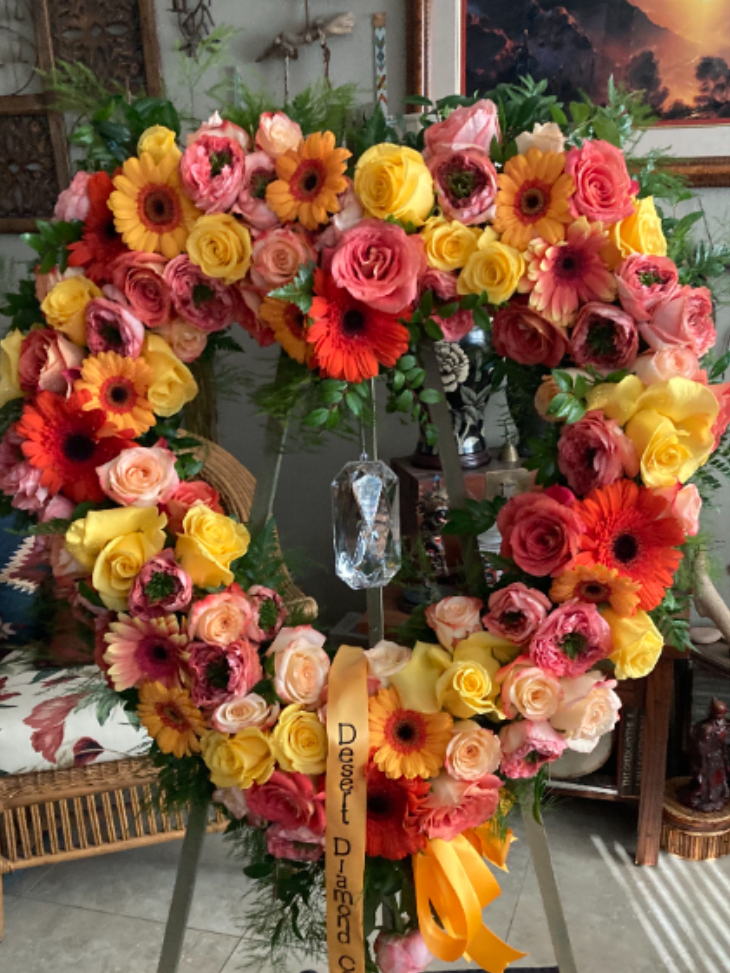 Heart-shaped standing wreath of pink, yellow, and orange roses and gerbera daisies on an easel with ribbon.