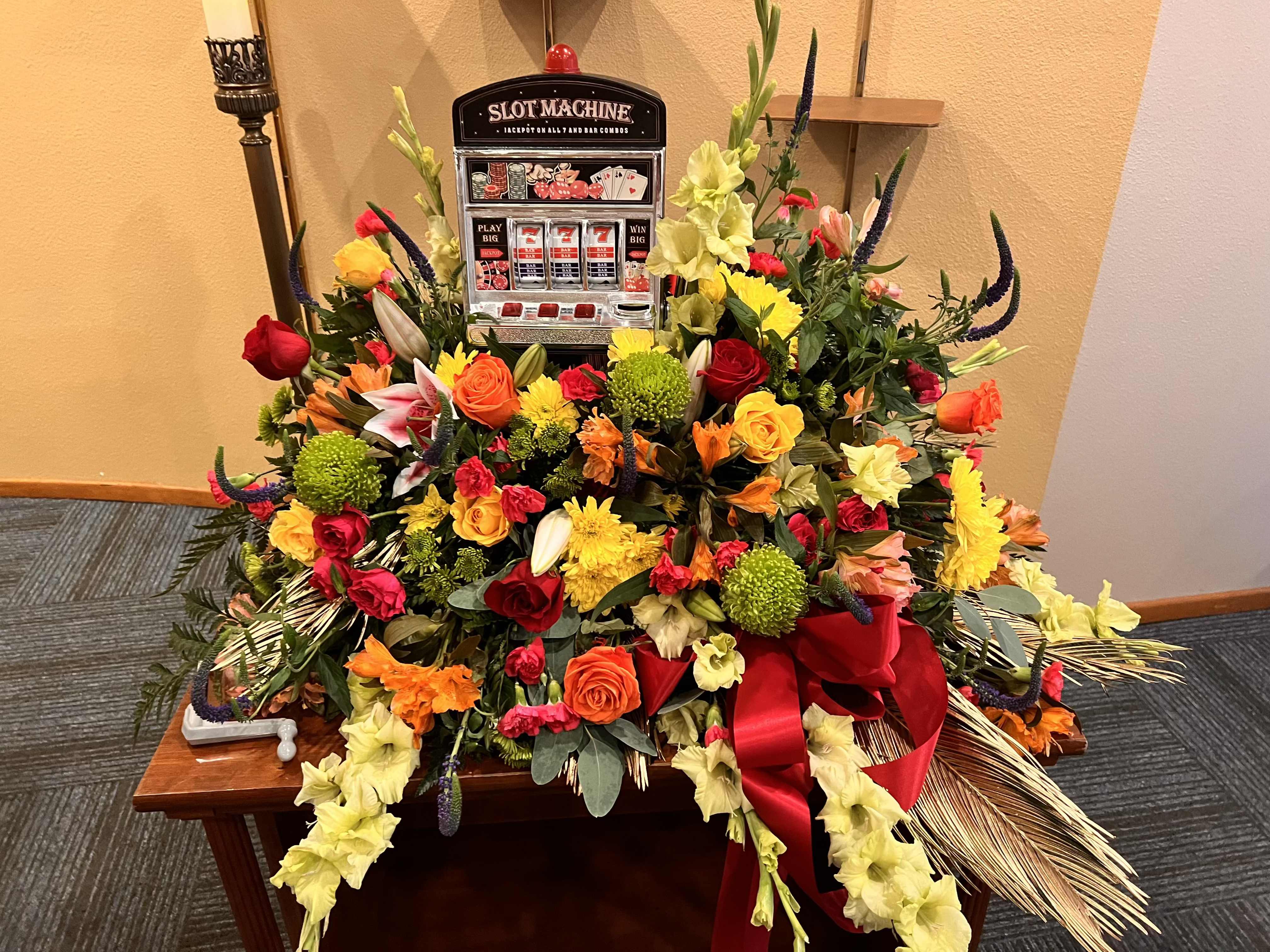 CASKET SPRAY - HOOT LOOT SLOT CASKET FLOWERS - REDS, GREENS, YELLOWS AND ORANGE FLOWERS AND FILLER FLOWERS WITH ASSORTED GREENS, AND A REAL SLOT MACHINE, ADORN THIS CASKET SPRAY, FOR THE CASINO LOVER.