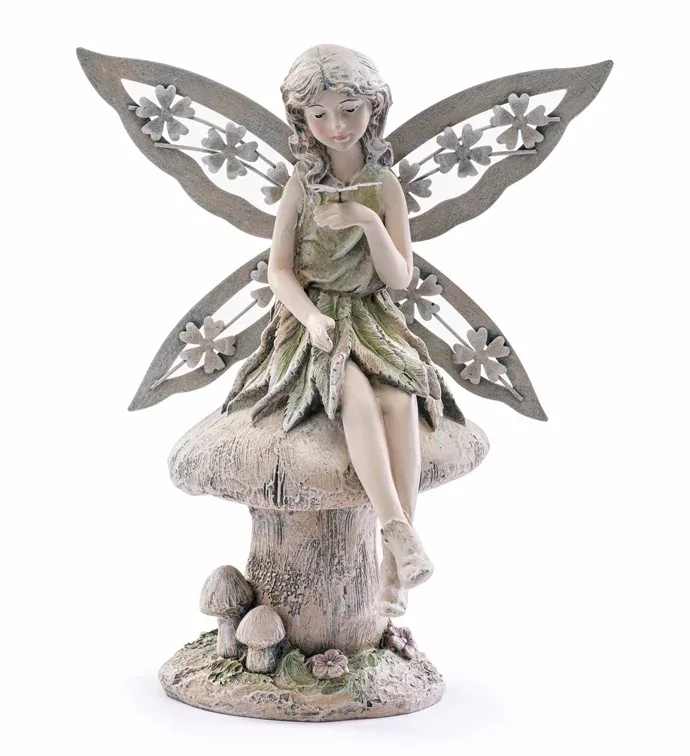 Fairy on Mushroom with Butterfly Figure  - A seated fairy atop a textured mushroom creates a storybook centerpiece that feels like a secret garden captured in stone - perfect as a garden-themed flower arrangement or a whimsical gift for a birthday, housewarming, or a thoughtful everyday surprise. The fairy-on-mushroom with butterfly figure sets a gentle, imaginative tone that will make the person you're thinking of feel seen and delighted by its quiet charm. The piece pairs muted whites, soft greens, and natural neutrals with sculpted flowers and smaller mushroom accents for an earthy, mossy look. The fairy's delicately detailed face, softly curled hair, and large cutout wings - accented with raised floral motifs - rest on a weathered stone-like mushroom base, making the decorative figurine a durable keepsake. Styled on its own or nestled into an arrangement, it brings texture and story to any display.  Ideal for someone who loves garden motifs, whimsical décor, or collectible accents, this figure works beautifully as a mantelpiece gift or a focal accent in a floral centerpiece. It's ready to place where it can be admired daily and pairs especially well with soft white blooms and greenery to echo its natural palette. 10.75 Inches in height and 7.25 Inches in width.