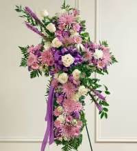 Florist Choice Faith Spray - A densely covered standing cross in soft pinks, purples and white offers a reverent tribute that speaks of faith and comfort - ideal as a funeral or memorial spray or for religious observances like Easter. The Florist Choice Faith Spray is a cross-shaped sympathy arrangement designed to console the person you're honoring and the family gathered to remember them.  This tall standing arrangement is formed with roses, chrysanthemums and complementary blooms in pink and purple, with clusters of white roses providing luminous accents. Spiky purple flowers reach upward from the upper left for a graceful, slightly asymmetrical movement, while a large purple ribbon bow and trailing streamers at the center reinforce the vertical line of the cross. Mounted on a standing easel, the piece presents a formal, church-appropriate silhouette with rich texture and soothing color contrast.  For services or graveside presentation, this spray conveys reverence and heartfelt remembrance; it's the kind of tribute that helps loved ones feel comforted and seen. The arrangement is delivered ready to display on the included easel - no assembly required - and our florists select the freshest blooms available to maintain color and form through the service.