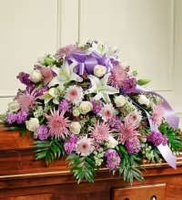 Florist Choice Lavender and Pink Casket Spray - Soft lavender and pale pink blooms create a peaceful, comforting tribute designed for memorial services and funerals. This lavender and pink casket spray lays low and elongated across a polished wooden casket, the arrangement composed to convey calm and respect so the person you're honoring feels remembered with tenderness.  White lilies and pale roses mingle with daisy-like chrysanthemums and clustered purple florals for a layered, textured look; touches of green foliage frame the pastel palette. A large lavender satin bow at the center and matching satin ribbon add a traditional, formal finish, while the cascading shape draws the eye along the length of the casket for a balanced, graceful presentation.  Ideal for family members or close friends seeking a refined, gentle expression of sympathy, this spray is arranged to sit securely on the casket and complements existing floral tributes. If you have specific requests for flower substitutions or ribbon personalization, please let us know when ordering so we can honor your wishes.