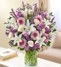 Violet Luxe Arrangement - A graceful mix of soft purples, creamy whites, and gentle pinks greets the eye-perfect for a sympathy arrangement, get well bouquet, or simply to let someone know you're thinking of them. The Violet Luxe arrangement pairs fragrant white lilies and ivory roses with lavender stock and pink-tinted chrysanthemums, accented by small purple daisy-like asters to create a soothing, uplifting presence the recipient will notice the moment it arrives.  The flowers are gathered in a clear glass cylinder vase that showcases straight green stems for a clean, classic look. This full, rounded design blends the lilies' elegant petals, the roses' timeless form, the stock's vertical texture, and the chrysanthemums' soft fullness into a balanced, traditional composition that suits both everyday gifting and more reflective moments.  Thoughtful and versatile, this arrangement makes the person you're sending it to feel comforted and seen-whether brightening a hospital room, offering condolences, or marking a quiet celebration. Arrives ready to display; change the water every two days and trim stems slightly for the longest vase life.