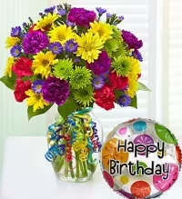 Timeless Celebration Arrangement - A burst of cheerful color greets the recipient: bright yellow daisies, verdant green button chrysanthemums, vivid purple asters, and bold red and magenta carnations are gathered into a rounded birthday bouquet that immediately reads as festive and joyful. As a birthday bouquet and celebratory arrangement, it's the kind of gift that makes the person you're thinking of feel noticed and cheered-perfect for birthdays, congratulations, or any upbeat celebration.  This mixed bouquet is arranged in a clear glass vase with multicolor curling ribbons cascading over the front for extra movement and party spirit, and it arrives with a coordinating round "Happy Birthday" mylar balloon printed with bright dots to complete the gift set. The arrangement's compact, rounded shape makes it easy to display on a table or desk; the mix of textures-from soft asters and daisies to the ruffled carnations-adds visual depth and lasting appeal.