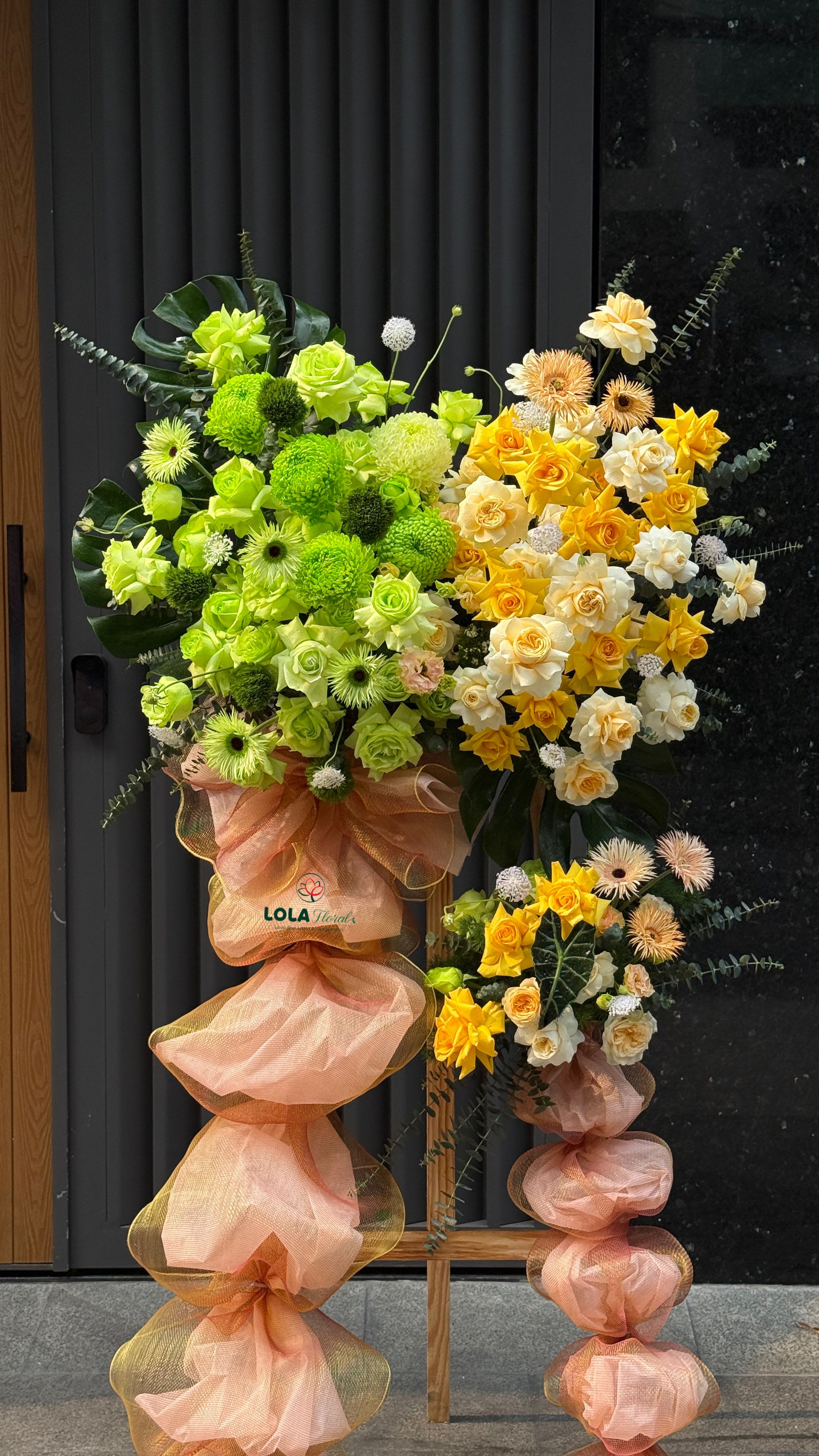 Grand Opening 2 - A bold curtain of chartreuse roses and green chrysanthemum pompons meets a sunlit spray of yellow and cream roses in this grand opening standing arrangement-perfect as a congratulatory display for a business opening, ribbon-cutting, or public celebration. The lively green, yellow, white, and peach palette reads joyful and energetic from across a room, giving the recipient the clear message: congratulations and well done.  Clusters of white ball-shaped blooms and spiky pale peach flowers add soft contrast and tactile interest, while gerbera daisies and mixed seasonal blooms fill the composition for a full, textured look. The arrangement is mounted on a tall wooden stand and finished with large peach mesh fabric wraps and cascading bows that reach the floor; a printed logo card is included for personalization and formal messaging.  Designed to command space and attention, this standing bouquet lets the honoree feel celebrated the moment they walk in. Ideal for storefronts, lobbies, or stage events-no watering required on arrival; professional handling keeps the display fresh for the duration of your event.