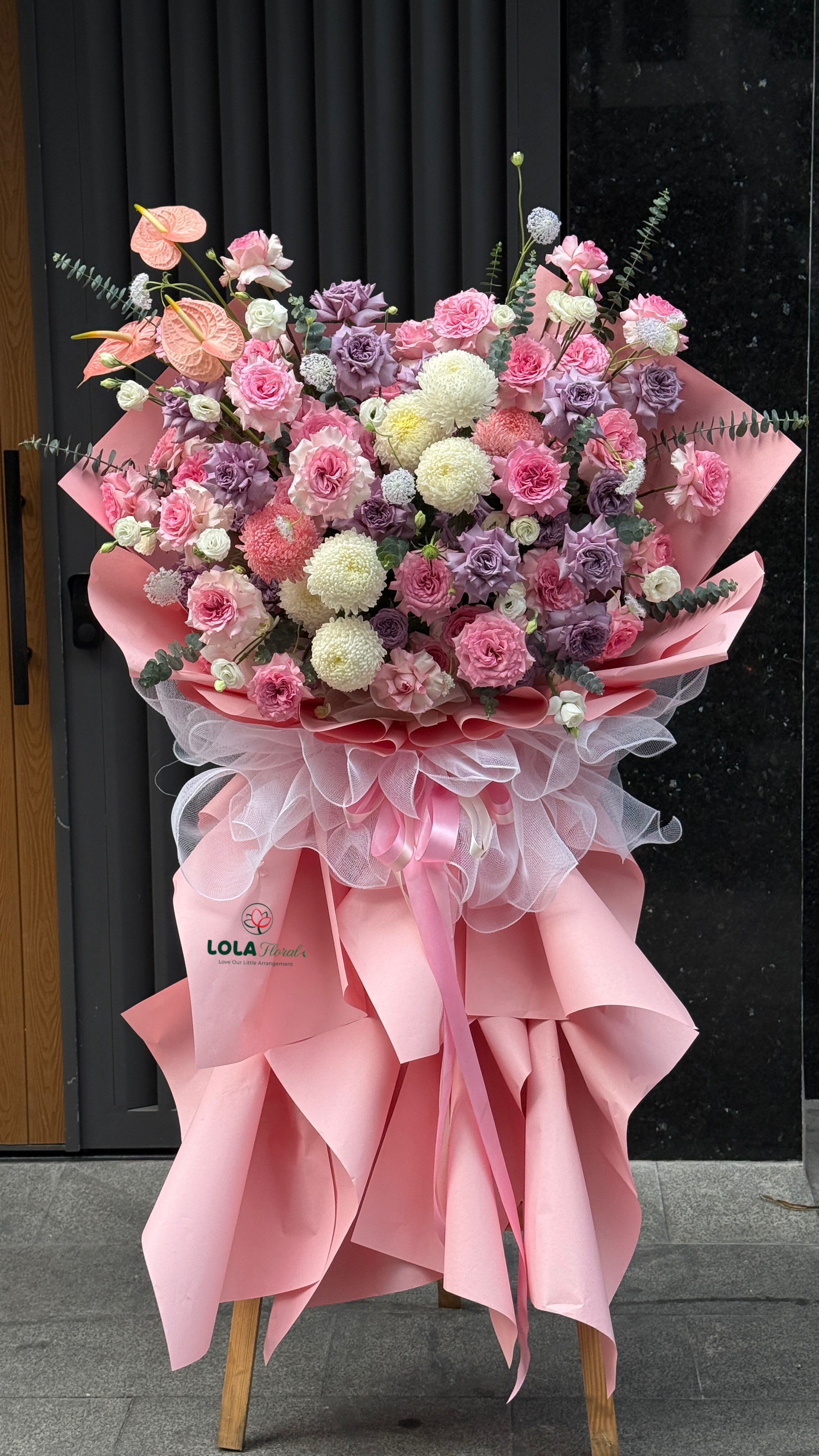 Grand Opening 3  - A lush cascade of pink, lavender, and white blooms makes this grand standing arrangement the perfect choice for a grand opening, congratulatory event, or a formal celebration like a wedding or anniversary. The mix of roses and spray roses with ruffled carnations gives a soft, romantic texture while pale pink anthuriums add a sculptural focal near the top - the kind of display that will make the recipient feel celebrated and honored.  This flower arrangement features a dense mass of roses, spray roses, carnations, and round white chrysanthemums, accented by a few pale pink anthuriums and tropical foliage for contrast. The bouquet is wrapped in layered blush pink paper and white mesh fabric, tied with flowing pink satin ribbons, and presented on a tall wooden stand for maximum presence. The pink, white, lavender, and purple palette reads feminine and festive, designed to command attention at any event.  Ideal for congratulating a business on a grand opening or marking milestone celebrations, this standing spray arrives ready to display and impress. Care is simple for event use-keep the base hydrated until the moment of placement and position away from direct heat; sizes and exact stem counts are crafted to fill the wooden stand and present a full, show-stopping silhouette.