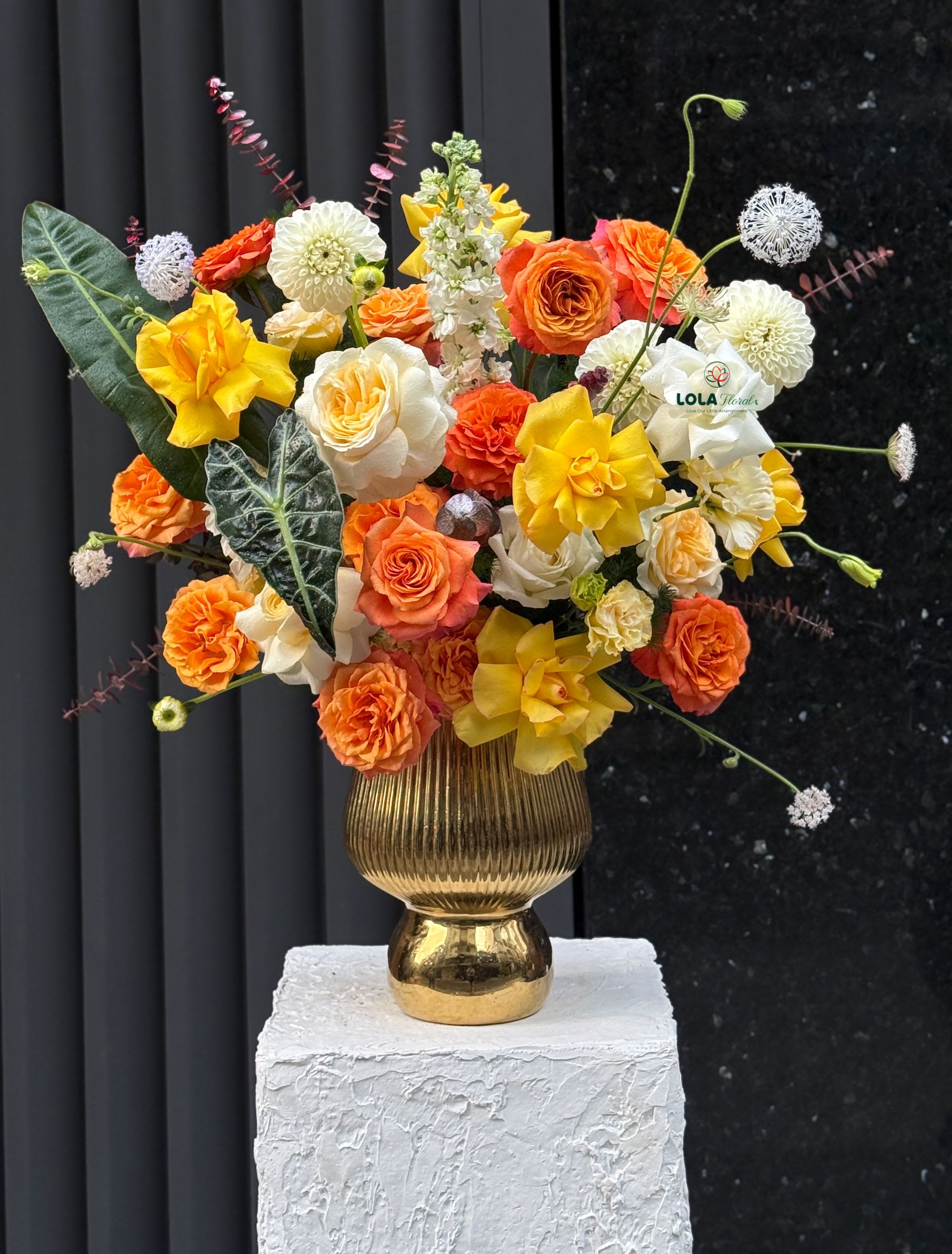 Orange Bloom - Warm, celebratory color takes center stage in this orange, yellow, and peach flower arrangement-a joyful gift-style bouquet perfect as a birthday bouquet, congratulations arrangement, or bright pick-me-up for someone special. The mix is designed to make the recipient feel truly celebrated: sunlit roses and spray roses sit prominently alongside round dahlia-style blooms, while upright white stock adds graceful height and a fresh vertical line that keeps the design modern and airy.  Arranged in a gold ribbed pedestal vase and shown on a white textured display block, the palette of orange, yellow, cream, and peach reads both luxe and approachable. Large garden-style roses anchor the silhouette, smaller spray roses and clustered filler blooms provide texture and movement, and the dahlia-like forms add a pleasing roundness. The overall size and loose, contemporary shape give this design presence without feeling stiff-it's the kind of arrangement that looks purposeful on a mantel, desk, or dining table.  Send this when you want her to feel noticed and cheered-ideal for milestone birthdays, promotions, or to say "well done" with warmth. The gold pedestal vase makes presentation effortless; light trimming of stems and fresh water will extend vase life and keep the composition looking its best for several days.