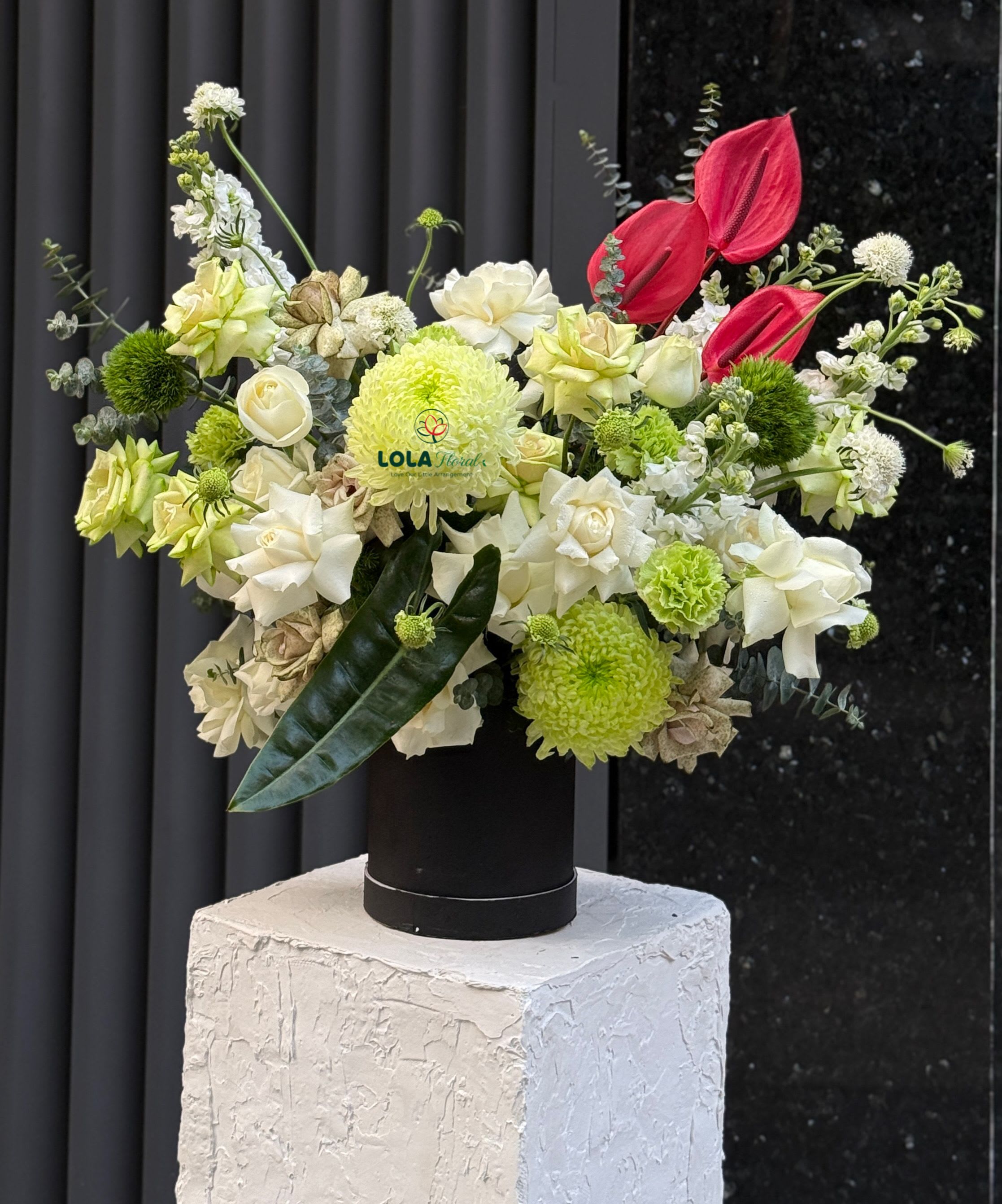 Green and White roses - A tall, sculptural flower arrangement built around ivory and pale green roses creates an immediate modern statement-perfect as a birthday bouquet, anniversary roses, or a contemporary celebration centerpiece. The green and white color story is punctuated by a few striking red tropical blooms that draw the eye, making the recipient feel celebrated and noticed the moment the arrangement arrives.  Creamy roses mingle with rounded green chrysanthemums, a single green carnation, pale hydrangea clusters and soft chrysanthemums for depth and texture, while tropical red accents slice through the neutrals for dramatic contrast. The stems are gathered in a sleek black cylindrical vase and presented on a rough-textured white pedestal to emphasize the arrangement's sculptural, gallery-ready profile.  This is the kind of gift that reads as thoughtful and stylish-ideal for someone who loves clean lines and surprising color pops. Sized as a tall, asymmetrical display, it's designed to command attention on an entry table or dining room console; keep water fresh and trim stems every few days to extend bloom life.