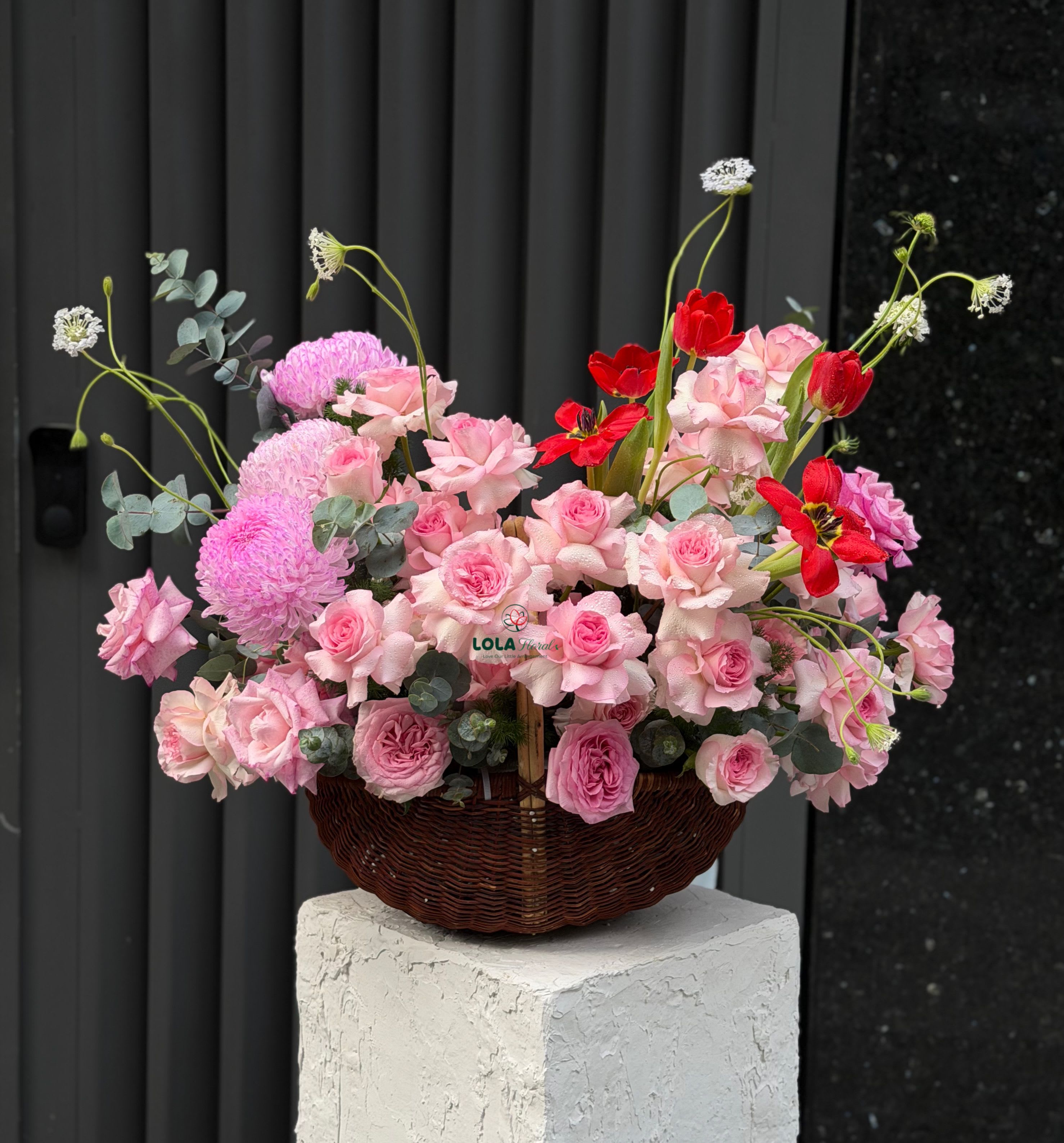 Pink Purple Basket - Dense clusters of pink roses in varying shades give this flower arrangement an immediate, romantic impact-perfect as a birthday bouquet, anniversary roses gift, or a loving "I'm thinking of you" surprise. The low, dark wicker basket grounds the design and makes the composition feel intimate and hand-delivered, while the red anemone-style blooms add bold contrast so the recipient will notice it from across the room.  Soft, round pink chrysanthemums add luscious volume and a cushiony texture against the smaller rose heads, and a few tall, delicate white blossoms rise above the main mass to introduce lightness and movement. The arrangement is designed low in a woven basket for easy placement on tabletops or mantels; the mixed pink, red, and white palette reads both celebratory and romantic without feeling fussy.  Ideal for anniversaries, Valentine's Day, or to celebrate a milestone birthday, this basket sends a message of affection and joy that will make the person you're thinking of feel truly seen. Care is simple: keep the floral foam moist and place the basket out of direct sunlight and away from heat to extend bloom life.