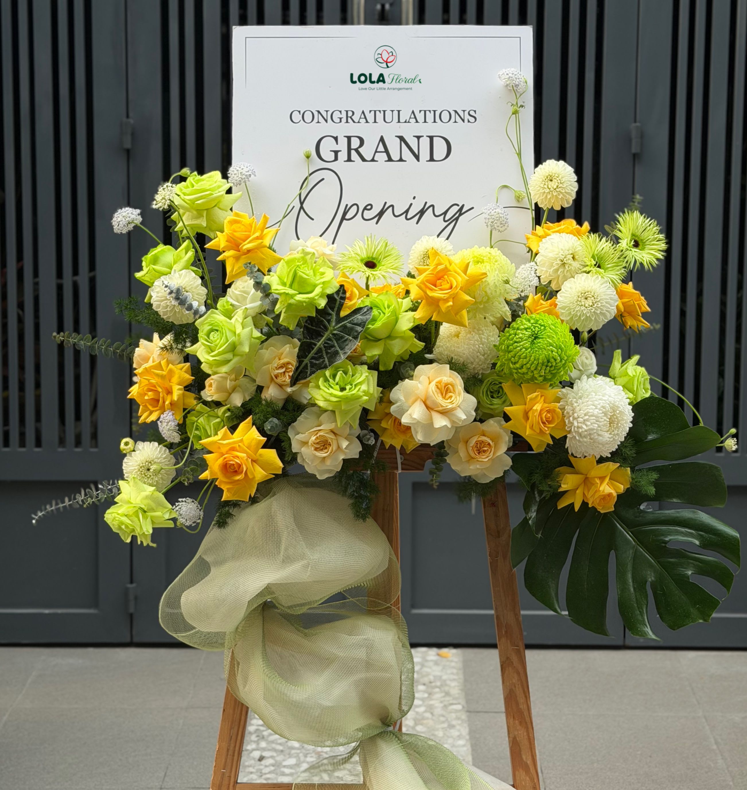 Grand opening  - Bright yellow and soft cream roses cluster with round white and green chrysanthemums to create a bold congratulatory display-perfect as a grand opening flower arrangement for a new shop, office ribbon-cutting, or milestone celebration. Mounted on a sturdy wooden easel and topped with a large printed sign, this standing display reads as an energetic, professional statement that will make the recipient feel celebrated and supported on their big day.  The arrangement sits densely at the base of the sign with yellow, cream and green roses, white and green chrysanthemum blooms, and small accent flowers for texture. A pale green sheer fabric drape trails down the easel for added vertical movement and airy softness, balancing the vibrant palette with subtle peach and green tones. Designed for corporate or large-scale events, the display arrives ready to set up on-site-an impressive visual send-off that draws attention and conveys warm congratulations.