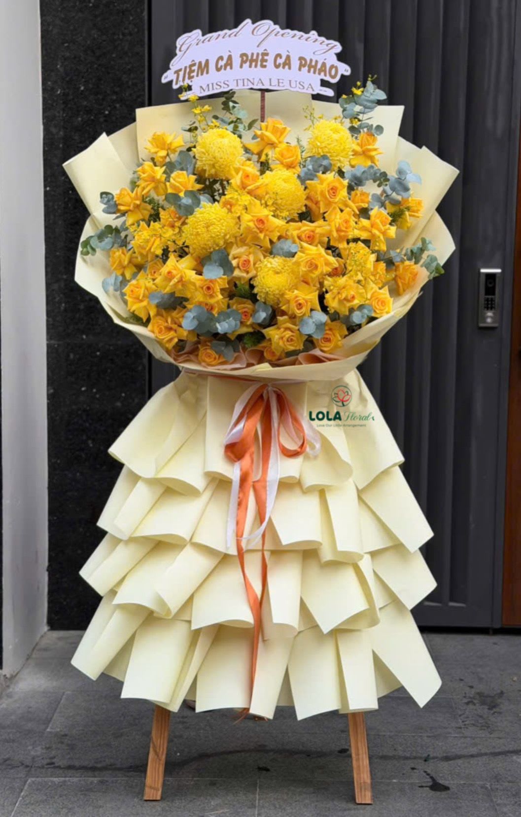 Blooming Success - Bright yellow roses and oversized round yellow chrysanthemums take center stage in this event bouquet-an attention-grabbing flowers for events perfect for a grand opening or congratulatory ceremony. Mounted on a tall wooden easel, the arrangement has the scale and vertical presence to mark an entrance or storefront so the person you're celebrating will feel the moment is truly special.  The design pairs dense clusters of chrysanthemums and roses in sunny yellow with accents of pink, white and orange peeking through, wrapped in layered cream paper that falls in structured ruffles like a skirt. A long orange-and-white ribbon flows from the center and a printed "Grand Opening" sign tops the piece, giving clear celebratory messaging. The easel adds height and stability for placement outdoors or inside a lobby.  Choose this for a new business launch, ribbon-cutting, or milestone ceremony to make the recipient feel celebrated and seen. The arrangement arrives ready to display on delivery; place on level ground or a sheltered spot if used outdoors and remove any packaging only after positioning to preserve the ruffled paper skirt.