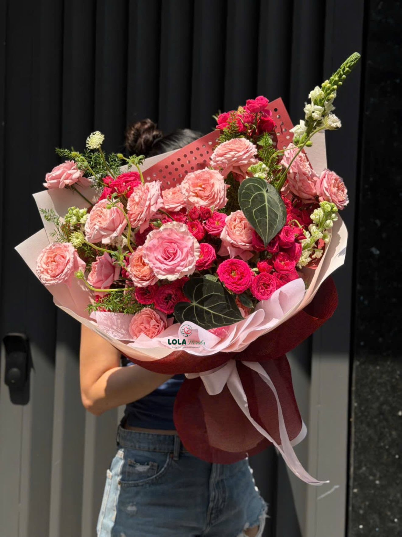 Pink Rose - Lush clusters of pink and red roses create an immediately romantic statement in this large hand-tied flower arrangement-perfect as an anniversary bouquet, Valentine's Day gift, or a heartfelt way to say "I adore you." The dense heads of roses are softened by smaller spray roses and scattered filler blooms so the person you're surprising will feel surrounded by color and texture; a few tall white snapdragons add vertical drama and a touch of surprise.  This bouquet blends shades of pink, deep red, white, and fresh green foliage for contrast and depth. Dark green, heart-shaped leaves and textured fillers punctuate the rose clusters, while the stems are wrapped in layered pink wrapping paper with a perforated pink insert, burgundy mesh wrap, and tied with white and burgundy ribbons. A branded bouquet sleeve finishes the presentation, giving the arrangement a generous, celebratory look.  Ideal for marking anniversaries, birthdays, or a meaningful romantic moment, this arrangement reads as both festive and intimate-she'll feel truly seen when these blooms arrive. It's offered in a large, hand-tied scale that fills a room and holds its shape; for best freshness, keep in cool water away from direct heat and place in a draft-free spot when displayed.
