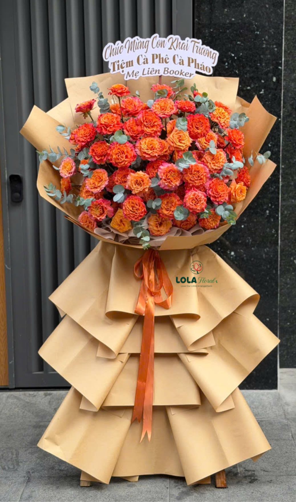 Timeless Beauty - A grand standing flower arrangement that reads as celebration from across the room, perfect as a congratulatory bouquet for graduations, grand openings, milestone birthdays, new baby welcomes, Mother's Day, anniversaries, or Easter-Timeless Beauty blends soft pinks and lavenders with bright orange accents to create an unforgettable, joyful display that will make the person you're honoring feel truly seen.  Dense, ruffled roses, carnations, and lisianthus form a plush textured surface punctuated by round white chrysanthemums and a few pale pink anthuriums for a distinctive tropical silhouette. Two presentation styles are available: a lush version wrapped in structured pink paper with voluminous white mesh, pink satin ribbon, and a wooden stand; or a bold orange-carnation variant set in tiered kraft paper with a tall pedestal, orange satin ribbon, silvery-gray filler stems, and a printed congratulatory sign. The scale is generous-designed to stand tall and command attention-so let us know your preferred stand, sign, or wrap when ordering; avoid direct heat and refresh any water source if provided for best longevity.