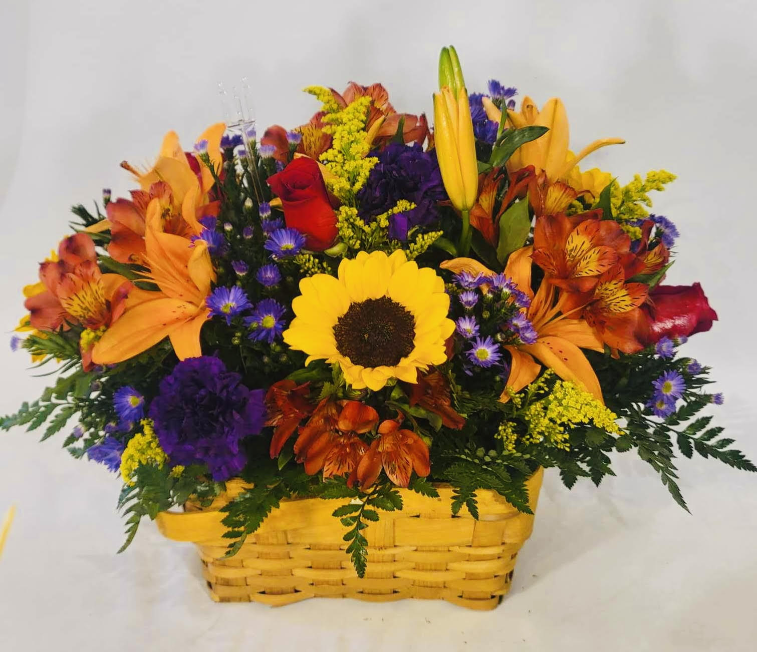  Basket of Blessings - A bright, sunlit sunflower takes center stage in this cheerful flower arrangement, making the Basket of Blessings a perfect birthday bouquet, get well gift, or Autumn centerpiece. The low, full composition in a yellow woven basket reads cozy and rustic-ideal for Mother's Day, Thanksgiving, housewarming, or simply to say "I'm thinking of you" with a warm, uplifting touch.  Orange Asiatic lilies and vivid red alstroemeria and roses surround the sunflower, while deep purple blooms and small purple asters weave contrast through the mix. Carnations and touches of yellow solidago add texture and fullness, creating a mixed-color palette of red, purple, orange and yellow that pops against the woven basket. The compact, generously filled design sits low for easy display on tables, desks, or counters.  Send this as a heartfelt anniversary surprise, a congratulations for graduation or retirement, or a just-because pick-me-up for a friend or grandparent. Its durable flowers and casual basket presentation make it equally at home for holiday gatherings or summer celebrations-order with confidence for same-week occasions and enjoy a gift that helps someone feel truly seen and celebrated.