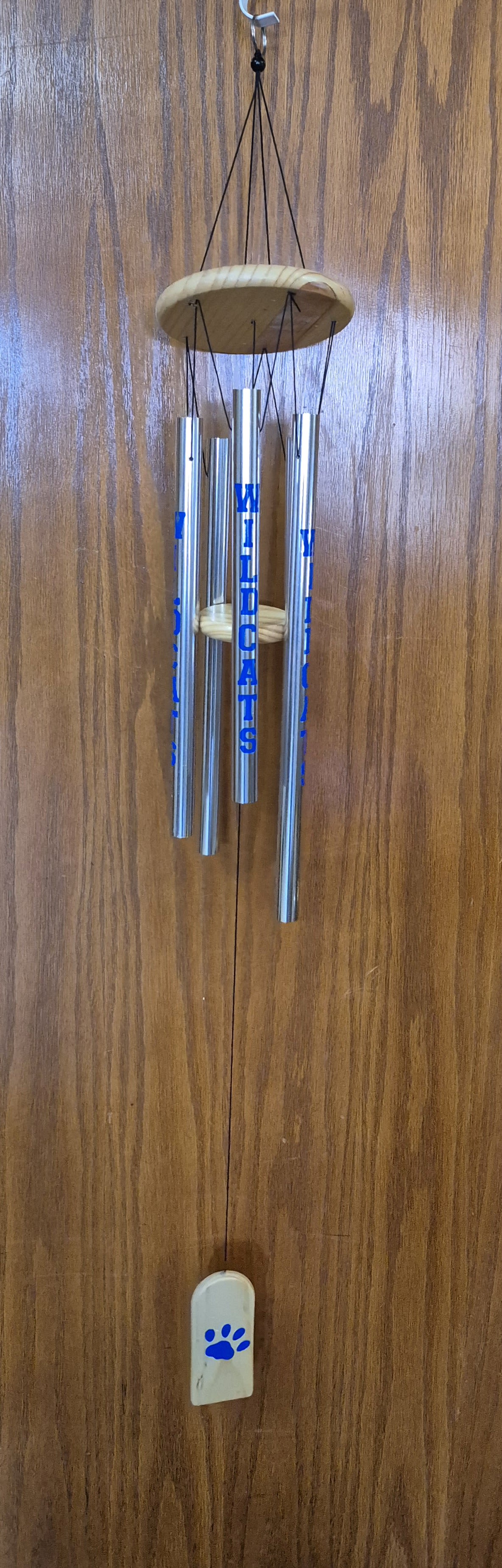  Kentucky "Wildcats" Wind Chime  - Bright silver tubes stamped with bold blue "WILDCATS" lettering catch the eye first, making this wind chime a spirited gift for a birthday, Father's Day, or to honor a favorite team at a summer tailgate. The gentle, music‑box tones from the hand‑tuned metal tubes offer a comforting, reflective sound that also suits sympathy and funeral displays-she'll feel remembered whenever the breeze stirs the wooden paw‑print sail.  Crafted with a light wood top disk, wooden clapper, and a matching wooden wind catcher printed with a blue paw print, the chime measures 44" from the top gathering ring to the bottom of the wind catcher and hangs easily from the included metal hook. The slim, neutral design works on porches, patios, or beside a memorial and complements the optional black metal easel stand for upright presentation (Deluxe price adds the stand).  Thoughtful for get-well wishes, Best Friends Day surprises, or to celebrate a boss who loves their alma mater, this wind chime sends a clear, personal message without words. Order with confidence for delivery year‑round; advise the recipient to hang it where breezes can move the sail for the best sound and longevity.