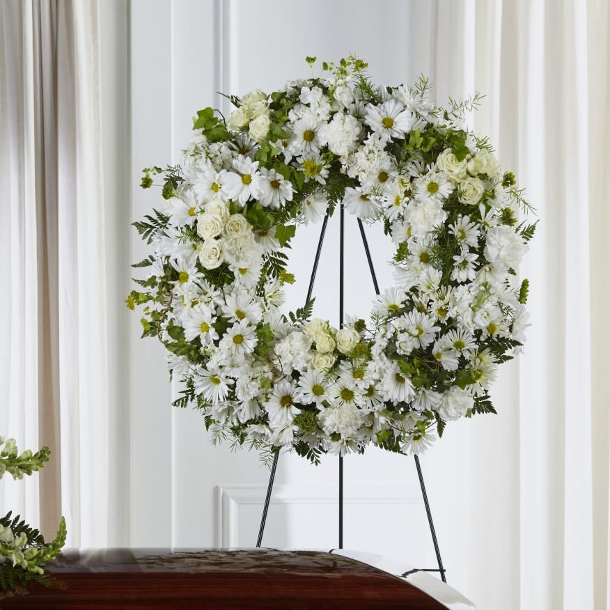 Pure Grace Wreath - A serene ring of white blooms makes an immediate, comforting impression-this standing wreath is a funeral arrangement built to express sympathy and quiet reverence. The Pure Grace Wreath is a circular standing wreath for a memorial service or graveside tribute, sending a message of peaceful remembrance to the person you're honoring.  Densely arranged white daisies with soft green centers sit alongside clustered white roses and cushion-like chrysanthemums to create a full, pillow-soft texture; small touches of green foliage add gentle contrast and depth. The wreath is mounted on a tall metal easel for prominent display, and is photographed beside a polished wooden casket to show its dignified scale and presence.  For families seeking a respectful way to say goodbye, this wreath offers a calm, uplifting visual that helps the recipient feel seen and comforted. Sized for standing presentation and delivered ready-to-display on the easel, it arrives prepared for immediate placement at the service.