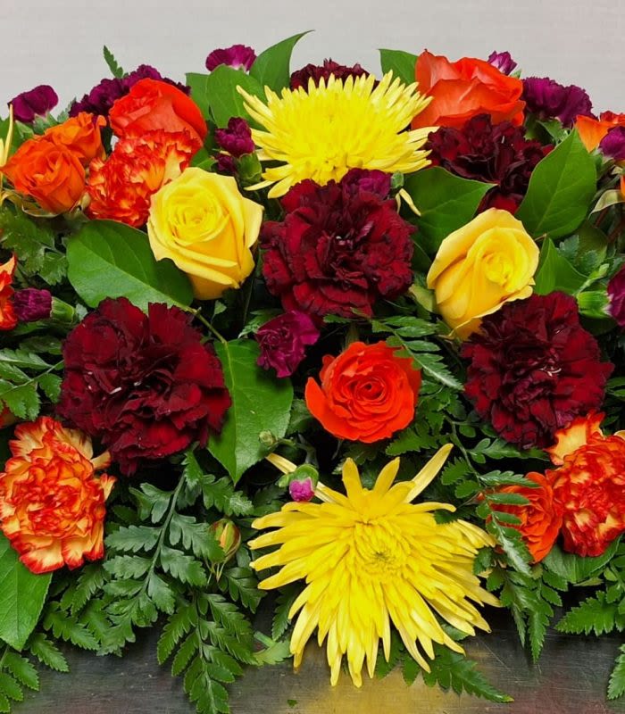 Autumn Skies Casket Spray - A low, elongated casket spray that brings warm autumn color and uplifting energy to a sympathy or funeral setting. This funeral flower arrangement places the product type (casket spray) and occasion (sympathy and funeral) up front while promising a comforting, celebratory presence for the person you're honoring.  Densely arranged blooms in red, yellow, orange and touches of purple create strong visual contrast and texture. Large yellow chrysanthemums (spider mums) punctuate clusters of deep red and bi-color orange carnations, while yellow and orange roses and lilies add rounded form and graceful height within the low profile. The design reads bright and energetic without being fussy, with a compact, elongated shape meant to rest across a casket.  Designed to comfort and to celebrate a life, this spray works well for families seeking a warm, uplifting tribute-especially in autumn months but appropriate year‑round. Arrives fully arranged and ready to place; stems are secured for stable presentation. If you need a custom size or specific color emphasis, contact us to personalize the spray for your service.