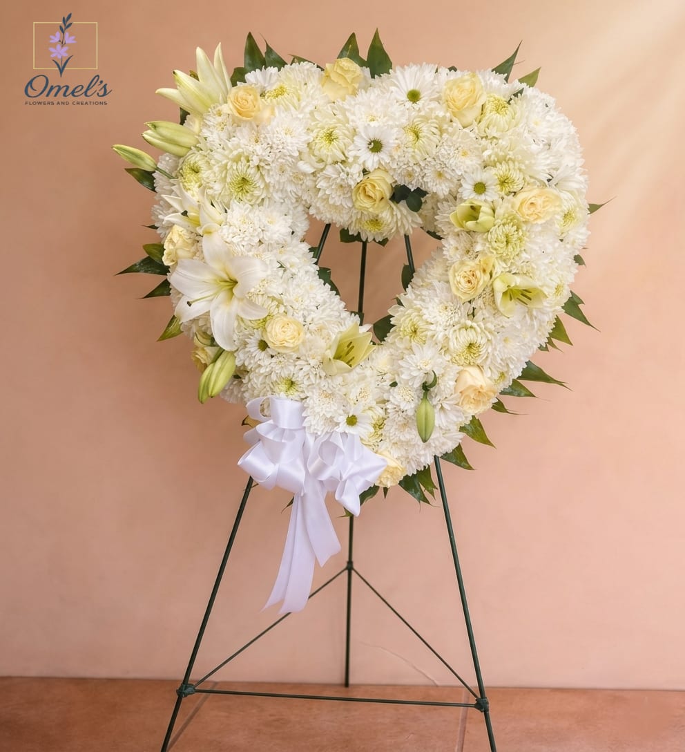 Hearted Everlight - A heart-shaped standing wreath of soft white chrysanthemums accented with pale yellow and cream roses and lilies-the kind of sympathy arrangement that reads as calm, reverent, and deeply considerate. Designed for memorials and funeral services, this heart arrangement sends a clear message of love and remembrance so the person you're honoring feels respectfully celebrated and gently remembered.  Densely packed chrysanthemums create a cushiony, full heart outline while clusters of soft yellow and peach-tinged roses and lilies add warm highlights and subtle vertical texture from a few unopened lily buds. The wreath is mounted on a tall metal easel and finished with a white satin ribbon bow with long trailing ends at the lower center, giving a formal, polished presentation appropriate for a service or graveside display.  Ideal for expressing sympathy, honoring a life, or offering condolences to family and friends, this standing heart wreath is sized for prominent display on the provided easel so it reads clearly from a distance. Care is simple-keep away from direct heat and replace water source foam as needed before service-to ensure the flowers look fresh and full for the occasion.