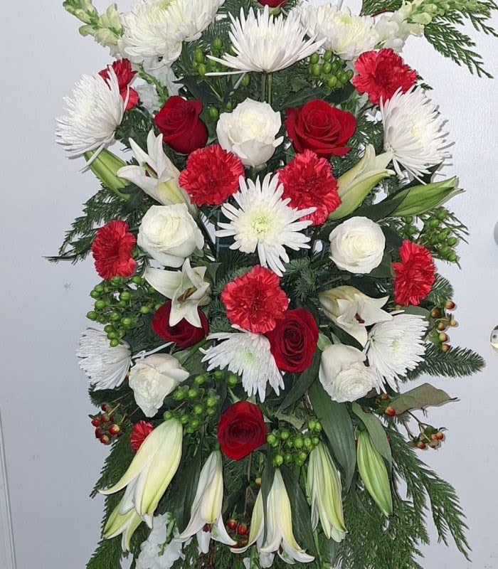 Blissful Beauty Standing Spray - Tall and solemn, this standing spray makes a dignified statement for sympathy and funeral services with a classic red, white, and green palette. The vertical arrangement of red roses and red carnations paired with white roses and trumpet-shaped white lilies creates a striking, respectful contrast that reads both formal and heartfelt-the kind of tribute that helps the person you're honoring feel remembered and respected.  Up close, the spray combines Asiatic lilies and white spider chrysanthemums with clusters of snapdragons, alstroemeria, and chrysanthemums for varied texture and layered depth. Evergreen foliage and small green-and-red berry clusters give the piece a slightly wintery, seasonal character while reinforcing a timeless, structured silhouette. The elongated form is designed to stand prominently at a service or graveside and conveys care through classic floral choices and thoughtful composition.  Ideal for memorial services or as a sympathy tribute to express condolences, this arrangement delivers a composed, comforting presence without words. Please let us know delivery details and any personalization requests so we can place the spray where it will be most meaningful; stems arrive ready for placement and benefit from fresh water at the service location.