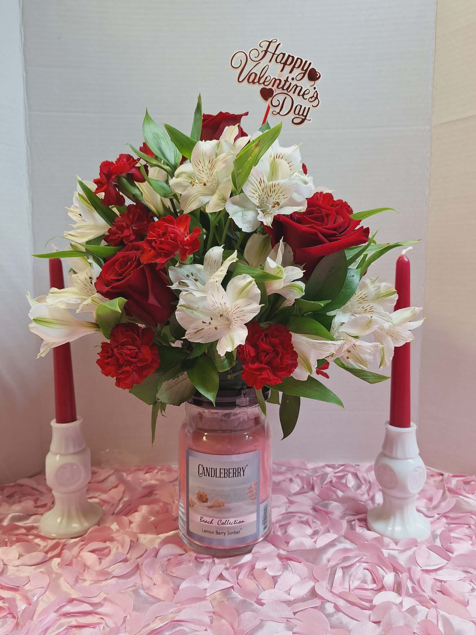 Candleberry Beach Collection Candle - A compact, romantic centerpiece that pairs fresh blooms with a scented glass jar candle-perfect as a Valentine's Day arrangement or for anniversaries and intimate celebrations. The rounded bouquet of red roses and red carnations contrasted with creamy white alstroemeria reads classic and heartfelt, while the scripted "Happy Valentine's Day" pick makes the romantic intent clear for the person you're thinking of.  The arrangement sits atop a glass jar candle and is styled on a pink rosette fabric covering, with matching red taper candles in white holders flanking the display for a warm, candlelit feel. Textures and colors-silky rose petals, ruffled carnations, and delicate alstroemeria-create visual depth and a lush, hand-tied look. The scent from the Candleberry Beach Collection candle adds a subtle, beachy fragrance to the floral notes without overwhelming the flowers.  Ideal for surprising a partner, marking an anniversary, or sending a birthday wish with romantic overtones, this piece arrives ready to display on a table or mantel. Standard care: keep flowers in fresh water, trim stems if needed, and never leave burning candles unattended; the arrangement is offered as shown with the candle.