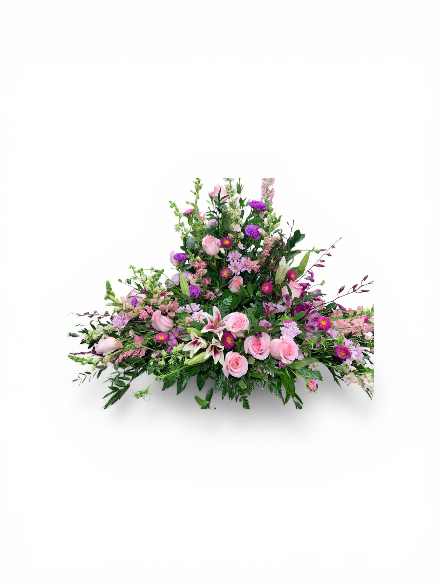 A Mothers Love Cremation Wreath  - A soft, elongated spray that speaks of tenderness and remembrance, this cremation wreath is designed for sympathy and funeral services where a gentle, comforting presence is needed. The arrangement's low, fanned triangular profile rises slightly in the center and tapers at the ends, giving it a graceful silhouette that honors a mother's memory while conveying calm and reverence.  Carefully clustered soft pink roses and star-shaped white lilies form the heart of the piece, while lavender chrysanthemums, purple snapdragons, and magenta alstroemeria add depth and quiet contrast across the length. Densely packed blooms create a lush, textured look with delicate pastel tones and occasional richer pops of color; the overall palette of pink, purple, white, and lavender reads both soothing and dignified. The design's horizontal format makes it ideal for placement on a casket, memorial table, or as a special tribute.  Made to comfort the person you're remembering, this wreath offers a meaningful way to say "we're thinking of you" to family and friends while honoring a mother's life. Size and handling details are available at checkout; to help the flowers arrive in their best condition, order in advance when possible so we can prepare and deliver them fresh for the service.