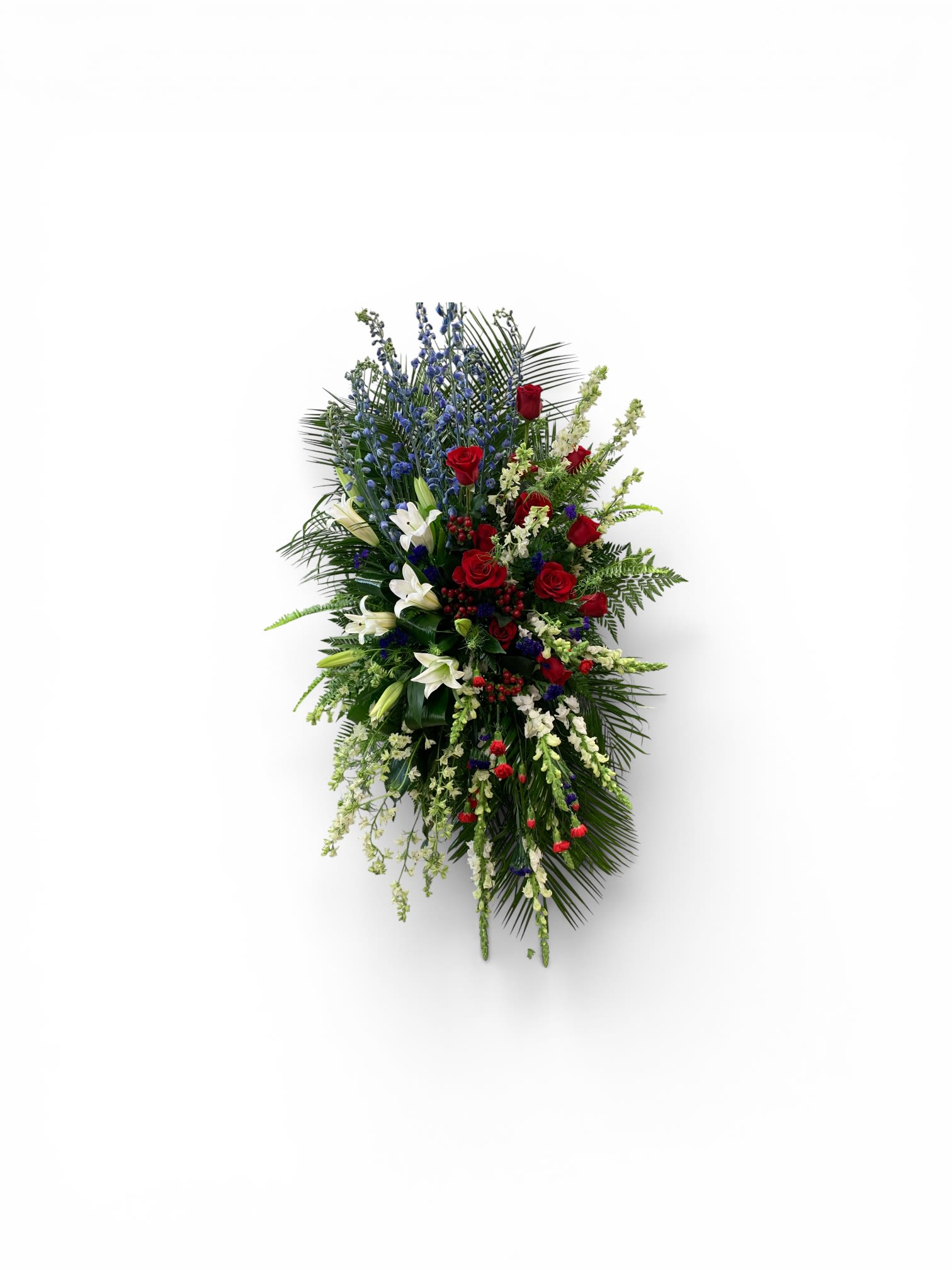 American Honor by Petal Perfection  - A dignified red, white, and blue funeral spray that conveys respectful remembrance and patriotic honor. This sympathy arrangement is a tall, elongated spray composed for memorial services and patriotic observances; the vertical, tapered silhouette gives it a formal, ceremonial presence that will stand beside a casket or at a graveside with quiet grace.  Clusters of deep red roses and fragrant white lilies are set against rising stems of blue delphinium and a lush backdrop of long fronds and mixed greenery. Smaller accent blooms in pale and red tones add texture along the edges and tips, creating layered depth and a balanced, dimensional look. The flowers are arranged with the blue stems reaching upward through the center while the lilies and roses form a richer middle cluster, preserving the classic spray shape.  Made to comfort the person you're honoring, this arrangement sends a message of respect and remembrance-an appropriate choice for memorials, Veterans' commemorations, or any time you need a solemn, patriotic tribute. Please contact us for size options and specific delivery needs so we can ensure the spray is arranged and presented exactly as required for the service.