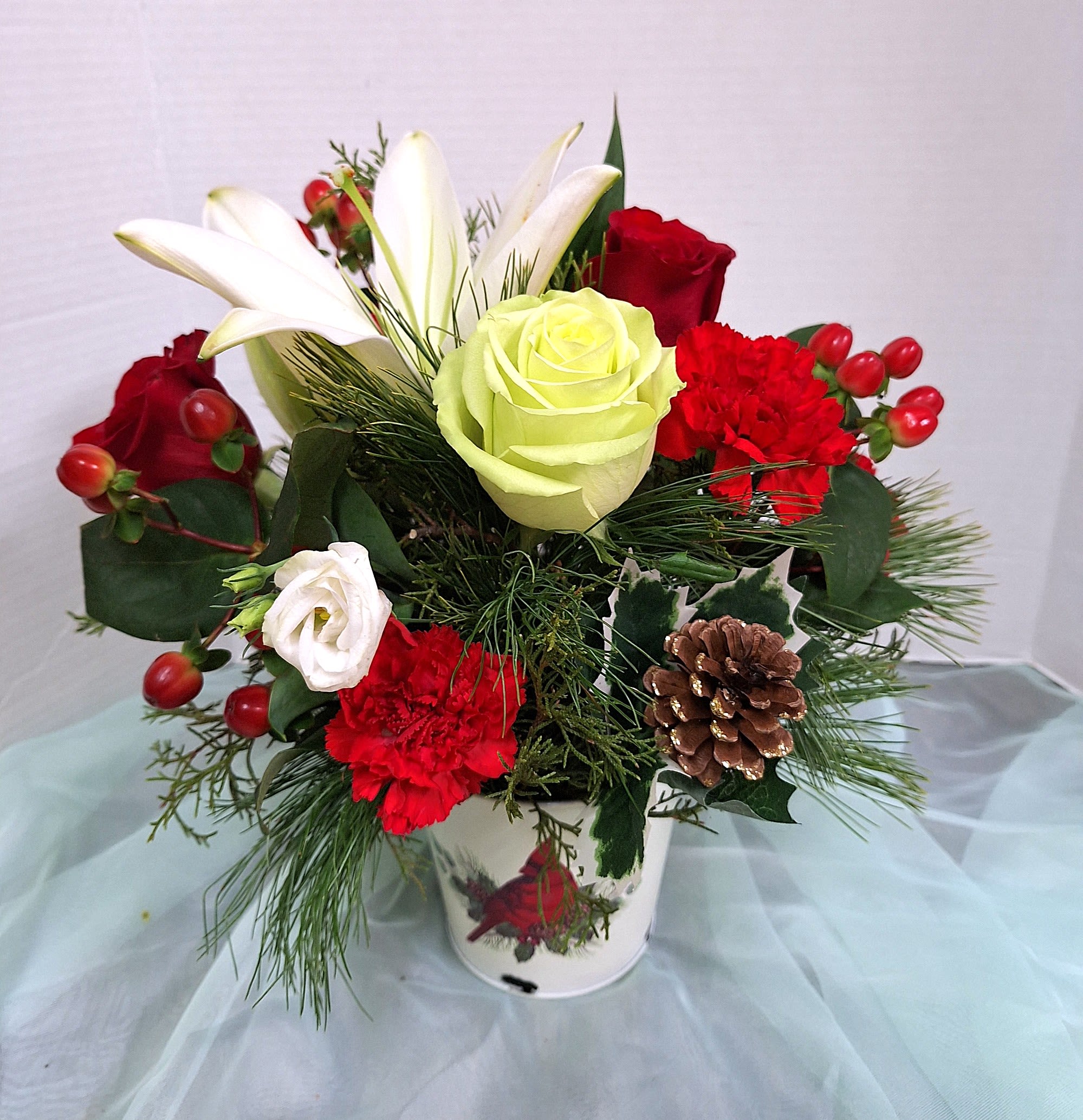 Comfort and Joy - A cheerful mix of red, white, and green designed to feel both festive and warmly personal - this holiday flower arrangement is a perfect Christmas or winter gift to brighten a mantle, table, or entryway. The arrangement reads as a holiday centerpiece but will also suit thank-you gestures or a thoughtful everyday surprise for someone who loves seasonal charm.  Clusters of white Asiatic lilies and pale green roses sit alongside classic red roses, red carnations, and delicate white lisianthus for a lush, textured look. Pine and winter greens frame red berries and a prominent pine cone, giving a woodland, seasonal touch. Everything is arranged in a decorative metal container printed with a red cardinal, adding a nostalgic holiday accent that still feels right any time in winter.  Give this to family or friends to convey warmth, comfort, and festive cheer - the kind of bouquet that makes someone feel genuinely seen during the holidays or on special winter occasions. Sized to work as a compact table accent or sidepiece, it arrives ready to display; keep stems in fresh water and trim as needed to extend bloom life.