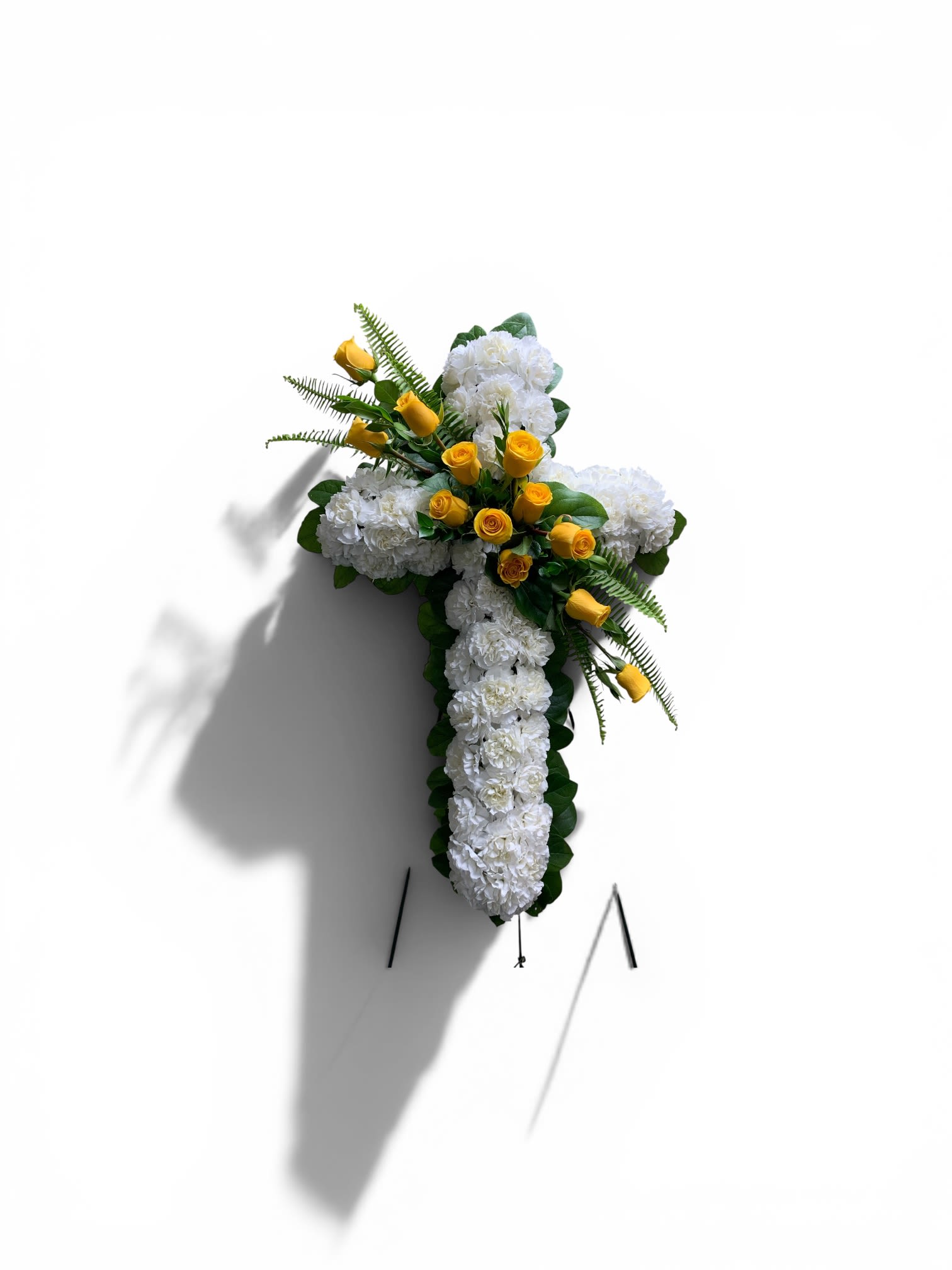 Bearer of the Cross  - A reverent standing cross crafted for sympathy and funeral services, this floral arrangement presents a dense field of white blooms shaped into a classic cross to honor a life and faith. The vertical presentation on a sturdy metal easel gives it a dignified, chapel-ready presence that reads as both comforting and respectful to those gathered.  White carnations and chrysanthemums form the solid cross, with darker green foliage subtly outlining the shape and adding depth to the design. A tasteful cluster of yellow roses and rosebuds accents one side, introducing a warm, hopeful contrast against the serene white; touches of lavender, peach and soft pink tones woven through the mix lend gentle nuance without distracting from the solemn form.  Designed to be displayed at memorial services or graveside, this arrangement communicates sympathy and reverence to the family and friends of the deceased. Mounted on the included metal easel for easy setup, it arrives ready to stand tall in a sanctuary or at a funeral home-no extra assembly required.