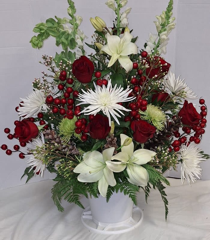 Eternal Elegance Funeral Basket - Deep red roses and creamy white lilies form the striking heart of this sympathy funeral basket, a floral statement that reads as both elegant and reverent. Designed as a funeral or sympathy arrangement, its tall, fan-like silhouette and seasonal hints - clusters of bright red berries and spiky white chrysanthemums - offer a respectful, comforting presence for a service or a loved one's home.  This arrangement pairs red roses, Asiatic lilies, and white chrysanthemums with green button-style blooms, snapdragon spikes, Bells of Ireland, asters, and accents of hypericum-like red berries for texture. The varied heights and shapes - from the upright Bells of Ireland and snapdragons to rounded roses and mums - create a structured, graceful profile. Flowers are gathered in a simple white plastic pot and presented on a round plastic stand, keeping attention on the rich red, white, and green palette and ensuring a stable display.  Appropriate for winter services or year-round remembrance, this basket expresses deep sympathy and thoughtful support for the person you're honoring. It arrives ready to display; to keep the arrangement looking its best, place it in a cool spot away from direct heat and replace water in the pot if provided according to florist care instructions.