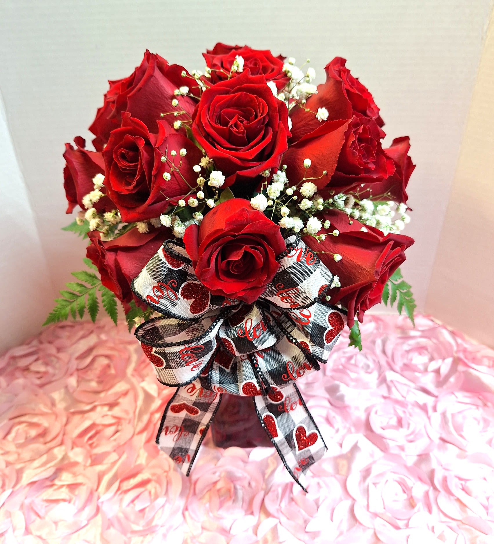 Loves Bounty - A compact cluster of rich red roses makes an immediate romantic statement-this anniversary flower arrangement and love-and-romance bouquet centers on deep red roses accented with delicate white filler blooms to brighten and sharpen the contrast. The design reads intimate and traditional, a thoughtful way to say "I love you" for anniversaries, Valentine's Day, or any moment you want the person you're thinking of to feel truly cherished.  The roses are arranged in a low, rounded grouping and presented in a simple clear glass vase that keeps attention on the vibrant petals. A large black-and-white checked ribbon bow, printed with red glitter hearts and the word "love," adds a playful, romantic accent at the front. Colors in the arrangement include red, white, and touches of pink alongside the bold black-and-white ribbon for visual interest.  Perfect for celebrating a milestone or surprising a partner with a meaningful romantic gesture, this compact arrangement suits bedside tables, dinner tables, or a special gift presentation. Care: keep vase water fresh and trim stems every few days to help the roses stay vibrant longer.