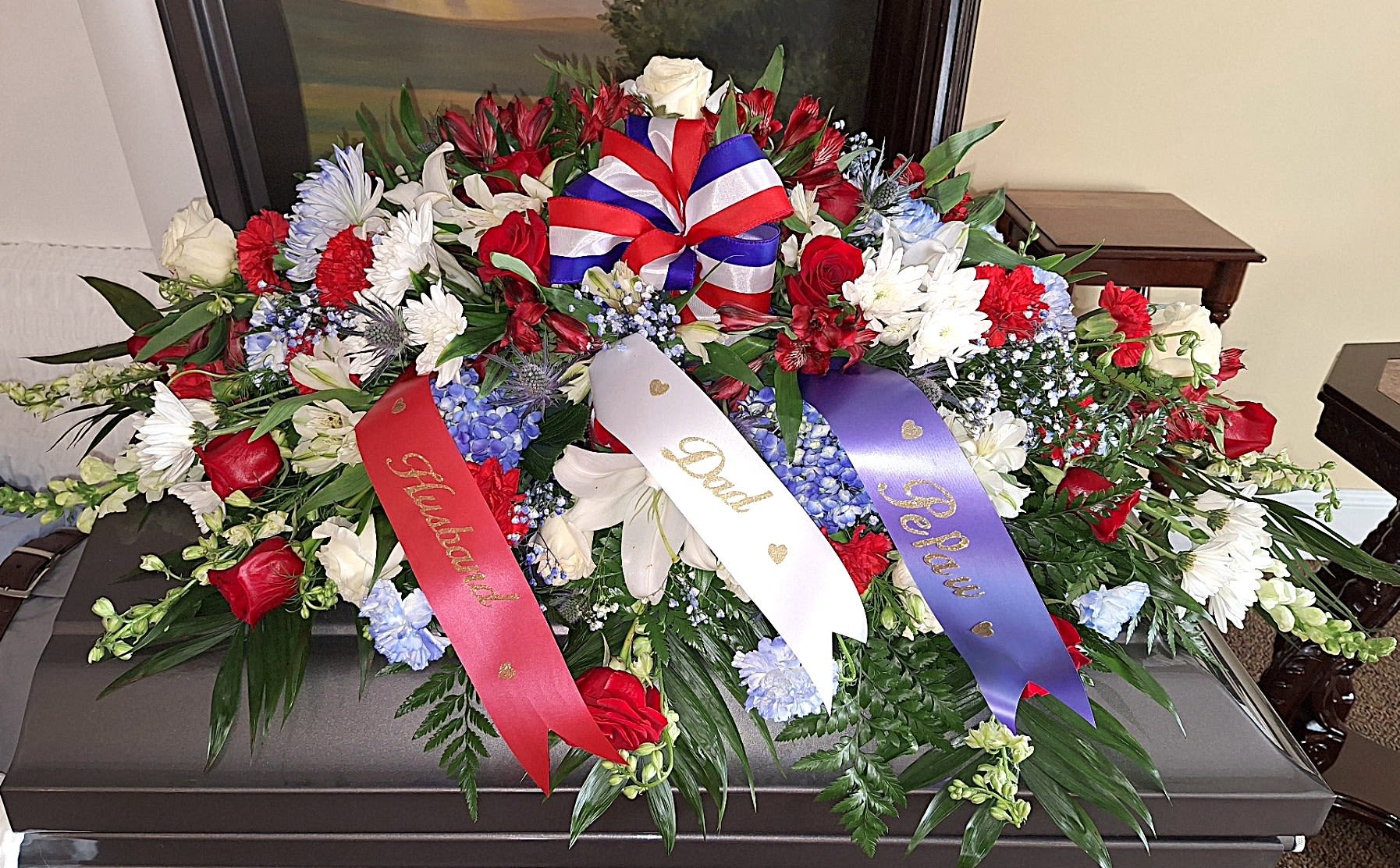Military Tribute Casket Spray - A dignified, full-length casket spray in a red, white, and blue palette that honors military service with quiet pride. This funeral casket spray for a sympathy or memorial service places red roses and carnations alongside white lilies and daisies, punctuated by blue hydrangeas and touches of purple delphinium and lisianthus to create a respectful, patriotic tribute the recipient will be remembered by.  The arrangement is long and gently domed to rest across the length of a closed casket, with dense clusters of blooms-roses, carnations, chrysanthemums, snapdragons, alstroemeria, asiatic lilies and hydrangeas-layered for texture and visual weight. Three satin tribute ribbons in red, white, and purple, each inscribed in gold script with family titles, drape across the spray and a red-white-and-blue bow crowns the center for a formal finishing touch.  Designed for a military or patriotic-themed memorial yet suitable for any sympathy service, this spray conveys honor, remembrance, and love on behalf of the family. For sizing and delivery arrangements or to personalize the ribbon wording, please contact us so we can ensure the tribute reflects the sentiment you wish to send.