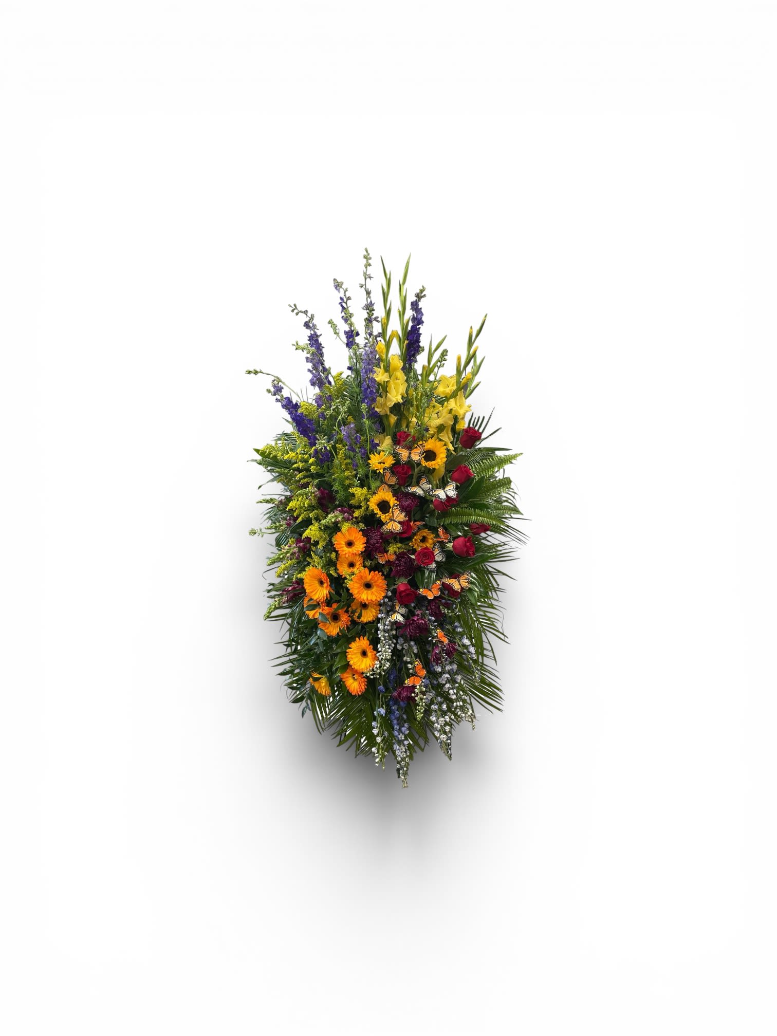 Monet’s Garden Spray by Petal Perfection  - A tall, uplifting spray that reads like a painter's palette-perfect for a sympathy or funeral tribute where you want to honor a life with color and dignity. This sympathy flower arrangement features the formal, vertical fan shape of a memorial spray while keeping the mood hopeful: bright yellow and orange blooms catch the eye, balanced by deeper red and purple tones for depth and reverence.  The composition includes clusters of gerbera daisies and other daisy-like blossoms concentrated toward the center and lower portion, with red roses and darker accents woven throughout to add weight and contrast. Spires of blue and yellow flowers reach upward against a lush green backdrop, giving the piece height and a garden-fresh texture. The mixed palette also carries soft hints of pink, lavender, peach and white among the chrysanthemums, carnations and delphinium, creating a layered, natural effect.  Designed to stand over a casket or rest on an easel at a memorial service, this spray offers a respectful, uplifting way to say farewell that will help the recipient's loved ones feel seen and remembered. Standard sizing fits most funeral displays; please contact us if you need a custom size or specific flower substitution for seasonal availability.