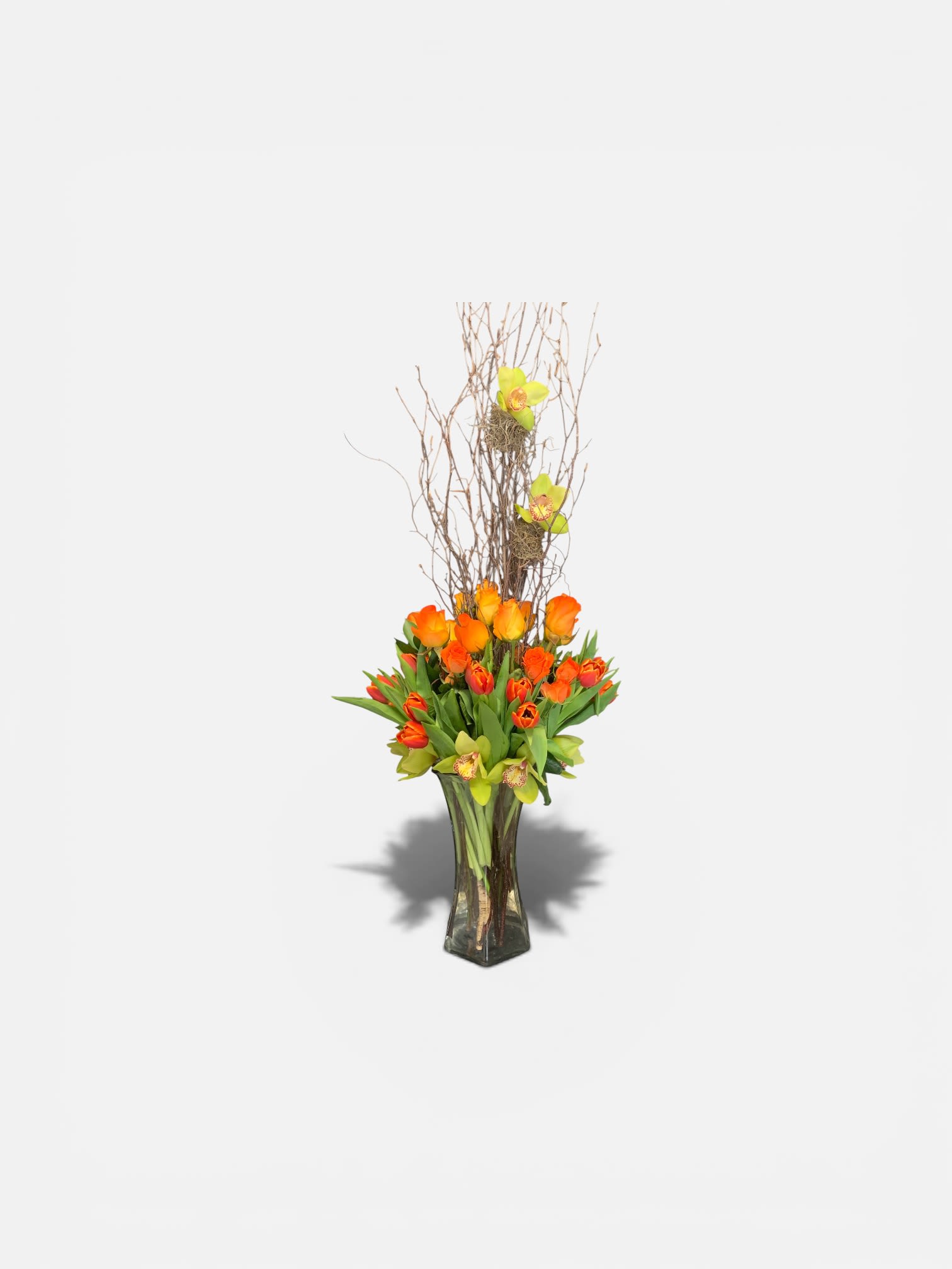 Orange Opulence by Petal Perfection  - A vibrant cluster of bright orange tulips crowns a tall clear glass vase in this contemporary spring bouquet - perfect as a birthday bouquet, Mother's Day gift, or a cheerful just-because arrangement. The bold orange and peach tones read joyful and energetic, while the vertical bare branches create a striking silhouette that gives the piece modern structure and presence for events like graduations, housewarmings, or seasonal Easter displays.  Peach roses and a few greenish-yellow cymbidium orchids tuck among the tulips and branches, adding exotic texture and soft color contrast that keeps the design from feeling one-note. The mix of orchids, roses, tulips and cymbidium orchid blooms creates a layered, tactile look: soft rose petals, the clean tulip faces, and the sculptural orchid centers. Display height comes from the tall clear glass vase and the upward-reaching branches, making this arrangement a strong focal point for tabletops, entryways, or office reception areas.  Designed to make the recipient feel celebrated and seen, this arrangement suits many occasions - anniversaries, congratulations, get well, sympathy, Thanksgiving or Christmas centerpieces, and everyday thinking-of-you moments. For best longevity, keep the vase topped with fresh water and trim stems slightly when refreshing; its tall profile also makes it ideal for gifting when you want to convey warmth, admiration, or bright seasonal cheer.