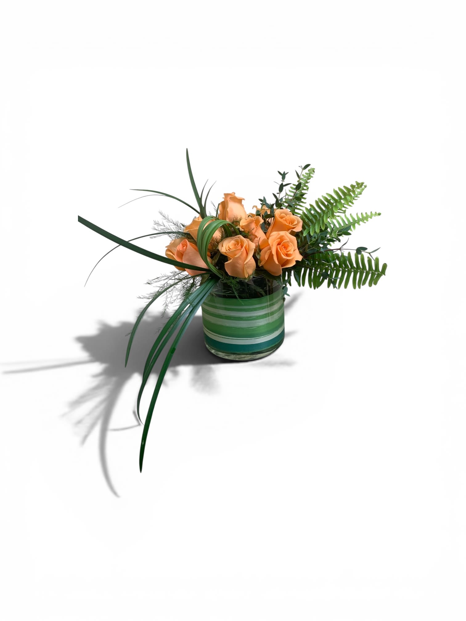 Peachy Keen by Petal Perfection  - Soft peach roses gathered in a compact cluster give this flower arrangement a gentle, modern charm-perfect as a peach rose bouquet for Mother's Day, an Easter centerpiece, or a fresh "thinking of you" gift. The low, round design keeps the focus on the warm, velvety blooms so the person you're sending it to will feel cared for without overwhelming their space.  Peachy Keen features peach roses set into a striped green glass vase with horizontal bands that echo the arrangement's clean lines. Arching blades of greenery and delicate fern fronds sweep outward to one side, creating an airy yet structured silhouette ideal for a small dining table, office desk, or bedside accent. Its compact scale makes it an easy housewarming, birthday, get well, or anniversary gift; it also fits beautifully into spring and summer celebrations without feeling seasonal.  Designed for effortless display, the low-profile vase arrives ready to place-no arranging required-and the arrangement's shape helps it stay tidy on tabletops and mantels. For best longevity, keep the water fresh and out of direct heat; this piece is a refined way to say thank you, congratulations, or simply that you care.