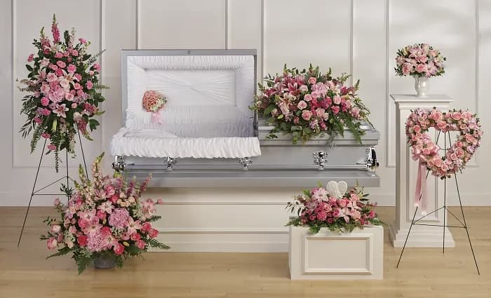 Petal Perfection’s All the Pink Sympathy Collection  - Soft pink roses and lilies drape gently across a casket in this tender sympathy flower arrangement set, creating a comforting centerpiece for a funeral or memorial service. The All the Pink sympathy collection pairs pink and white blooms with verdant greenery to convey warmth and remembrance-the kind of floral display that helps the person you're honoring feel seen and cherished.  This collection includes a full casket spray of pink roses, lilies, carnations, and snapdragons, plus matching standing sprays on metal easels, a heart-shaped wreath, low front arrangements, and a rounded bouquet arranged in a white urn-style vase. The palette of soft pink, white, and green is carried through each piece for a cohesive, elegant presentation; hard goods shown include a silver casket, metal easels, a white pedestal, a white rectangular container, a white urn vase, and an angel figurine to complement the arrangements.  Appropriate for a formal funeral or intimate memorial, these pieces can be ordered together for a unified display or selected individually to suit your needs. Arrangements arrive ready to place on casket or easel-simple care is limited to keeping flowers in fresh water and sheltered from direct heat to maintain their gentle color and form during the service.