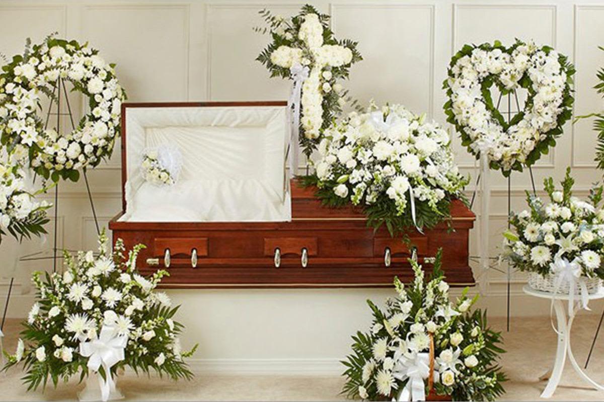 Petal Perfection’s All things Remembered  - Pure white blooms arranged with generous green foliage create a calm, reverent display for a sympathy or funeral setting. This flower arrangement group centers on classic lilies, roses, chrysanthemums, and carnations-presented as a casket spray, floor pieces, standing wreaths (including a circular and a heart shape), and a floral cross on a metal easel-so the person you're remembering will be honored with a cohesive, dignified presentation.  The arrangements feature textured white petals against abundant green leaves for a soothing, timeless palette; formal white ribbons and bows add quiet elegance. Hard goods include a wooden casket with a pale interior for context, white pedestal tables, white ceramic vases, and a white wicker basket where appropriate, giving each piece structure and a refined finish. Pieces are sold individually or together as a coordinated group to suit the service size.  Choose individual pieces to personalize a tribute or select the full grouping for a unified, respectful display that helps family and friends feel comforted and seen. Allow florists time to source seasonal white varieties and to secure requested hard goods; small care notes-keep flowers cool and out of direct sun-help them look their best through the service.