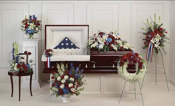 Petal Perfection’s American Duty - A formal, patriotic tribute fashioned in red, white and blue to honor military service or a life lived with devotion. This funeral flower arrangement collection centers on a casket spray that frames a folded American flag, with matching standing sprays, wreaths, and urn-style pieces arranged for a memorial or service. Designed for sympathy and funeral use, the coordinated display reads solemn and respectful-exactly the presence the person you're remembering deserves.  White lilies and carnations provide a graceful, fragrant foundation while vivid red blooms and blue delphinium and hydrangeas add striking, dignified color. Chrysanthemums and additional carnations fill the compositions for texture and lasting formality; pieces come presented on metal easel stands, a wreath stand, and a pedestal column, with ceramic urn vases, a small wooden table, ribbons, and an included picture frame available to complete the display. Available as a full coordinated set or ordered individually, each element is sized for venue presentation and designed to hold its shape throughout the service.