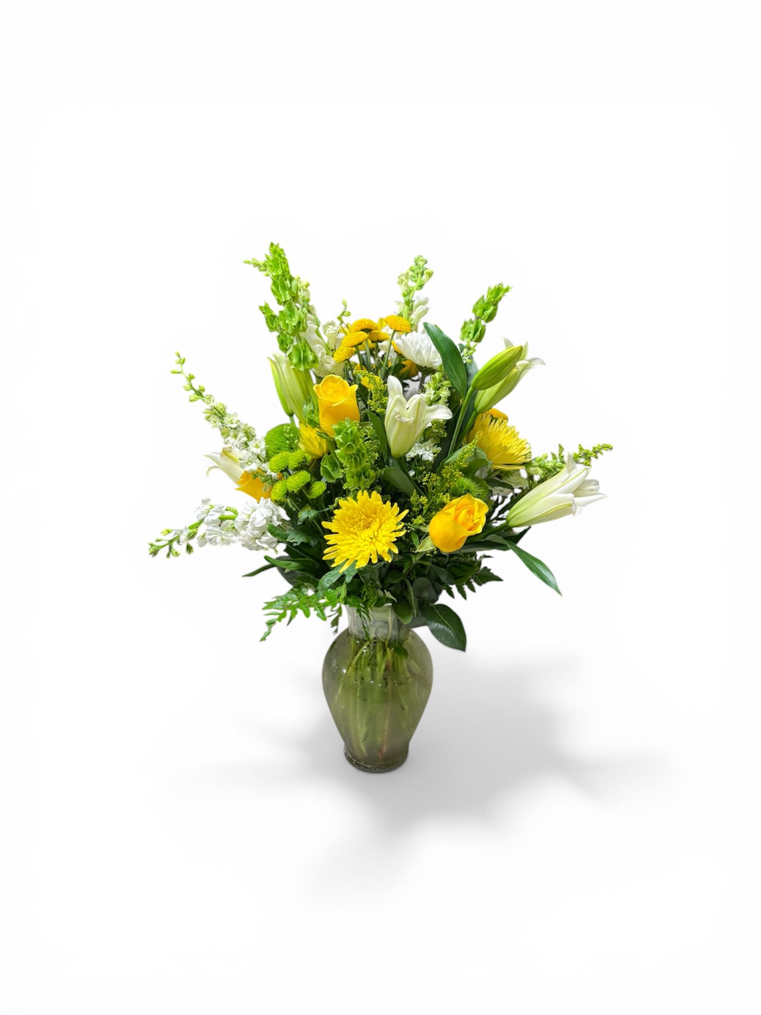 Petal Perfection’s Lemon Lime - Sunlit yellow roses and cheerful gerbera-like daisies meet crisp white lilies in a Lemon Lime flower arrangement designed to brighten any room - perfect as a birthday bouquet, Mother's Day surprise, get well gift, or an uplifting everyday gesture. The tall, airy silhouette makes this flower arrangement especially fitting for spring and Easter décor while remaining joyful and versatile for summer, anniversaries, new baby celebrations, or simply saying "I'm thinking of you."  This composition pairs yellow, green, and white blooms with vertical Bells of Ireland to add elegant height and structure, arranged in a green-tinted glass vase for a fresh, modern look. The mix of textured petals and upright greens creates visual movement and a light, hopeful feel; the lilies add fragrance while the roses and daisy-like flowers deliver long-lasting color. Sized to stand out on a mantle or brighten a bedside table, Lemon Lime is an uplifting choice for housewarmings, graduations, sympathy, or holiday gatherings - and it's easy to care for with regular water changes and fresh cuts to the stems for longer enjoyment.